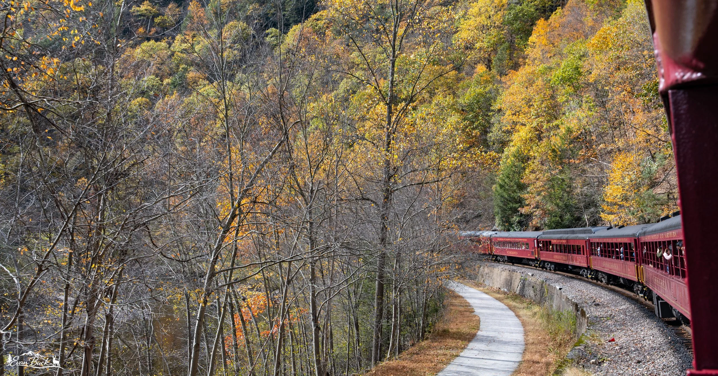 A red vintage train travels along a curved track through a forest with autumn foliage. The scene is viewed from a train car window, partially visible on the right.