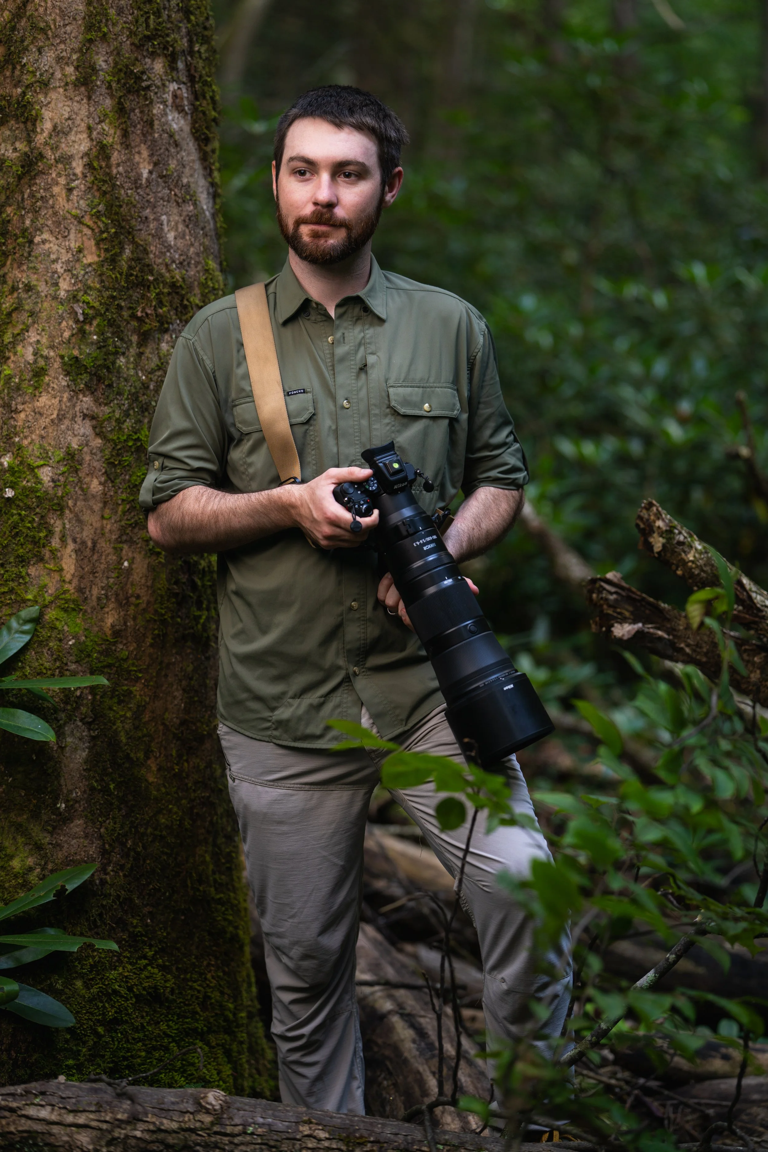 Evan Buck of Evan Buck Photography standing in a lush green forest, holding a professional camera with a large telephoto lens, wearing an olive green shirt and light-colored pants.