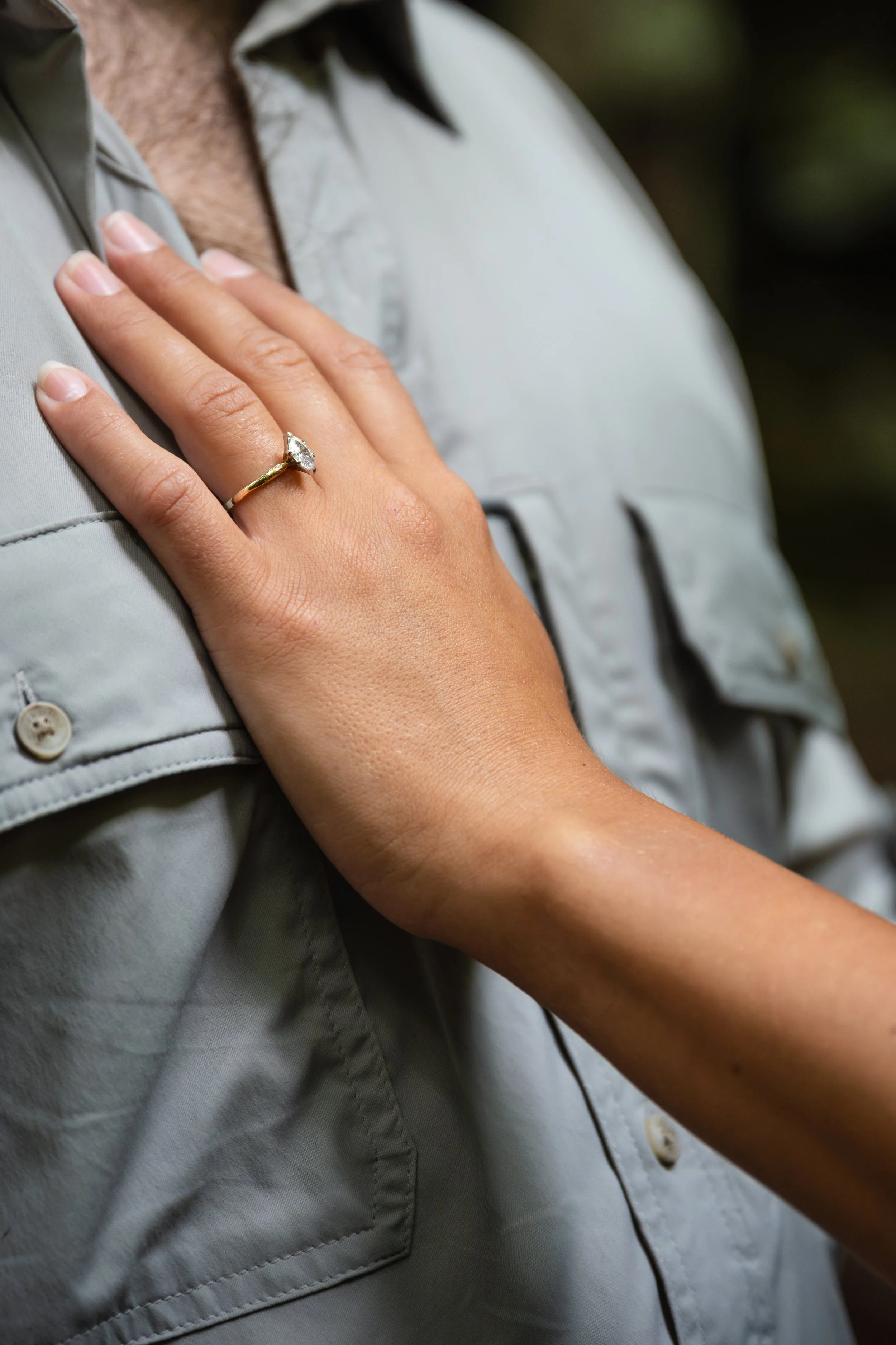 Close-up of a person's left hand resting on their chest, wearing a gold engagement ring with a diamond, and dressed in a light grey shirt.