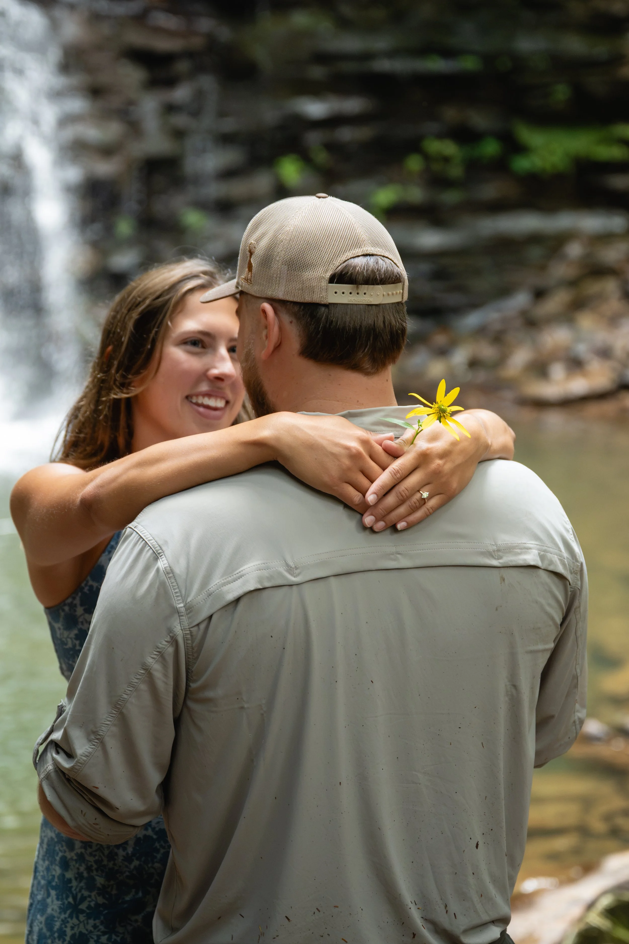 A young woman and man embracing outdoors near a waterfall, with the woman smiling and holding a yellow flower, and the man wearing a beige cap.