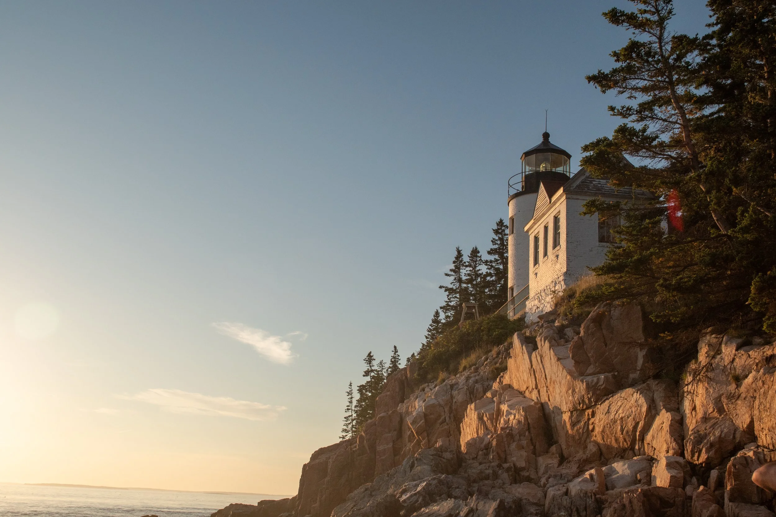 A lighthouse on a rocky cliff at sunset with trees and a clear sky.