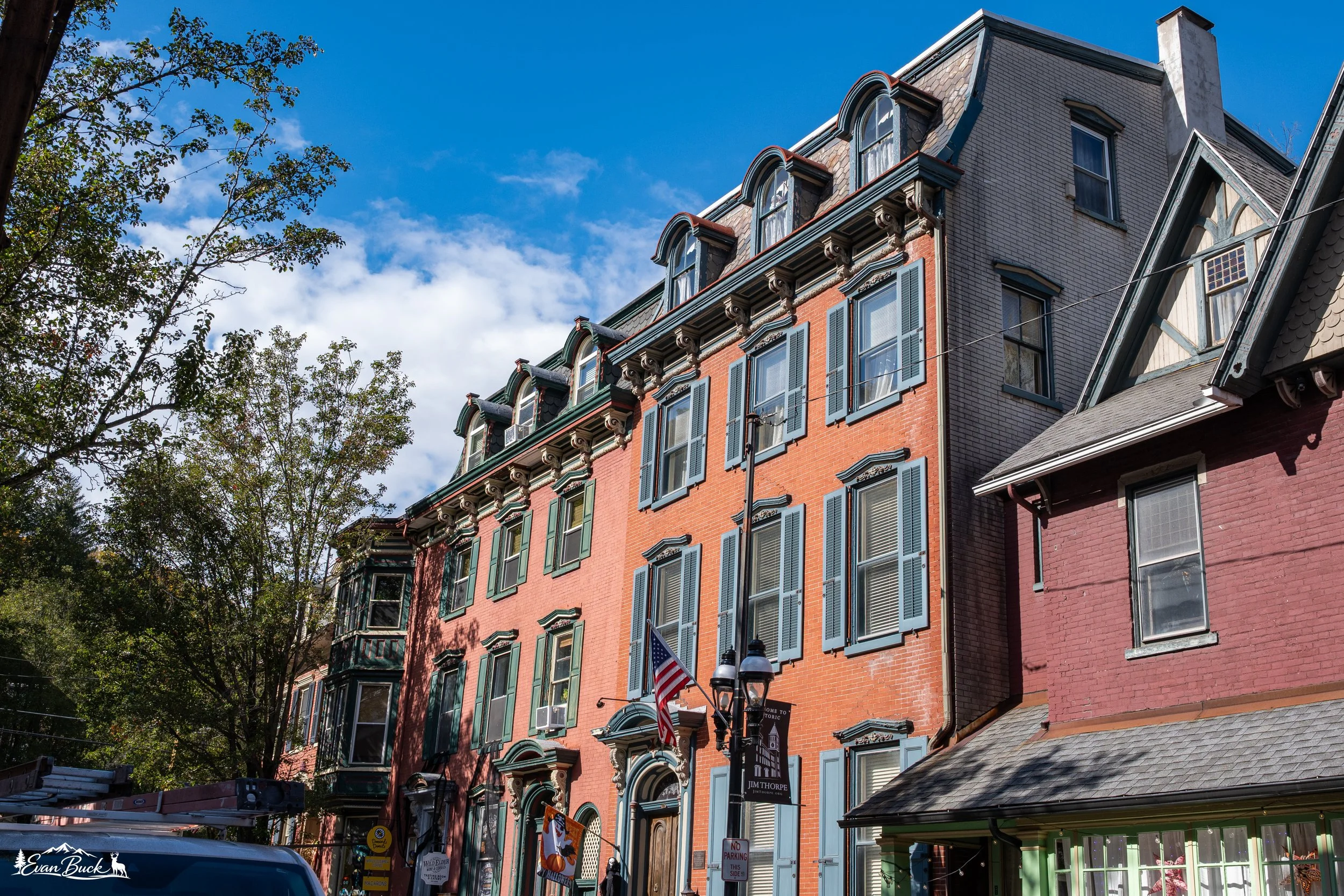 A row of old Victorian-style buildings with colorful facades on a sunny day with a blue sky and some clouds. There are trees on the left and a street with parked cars in front.