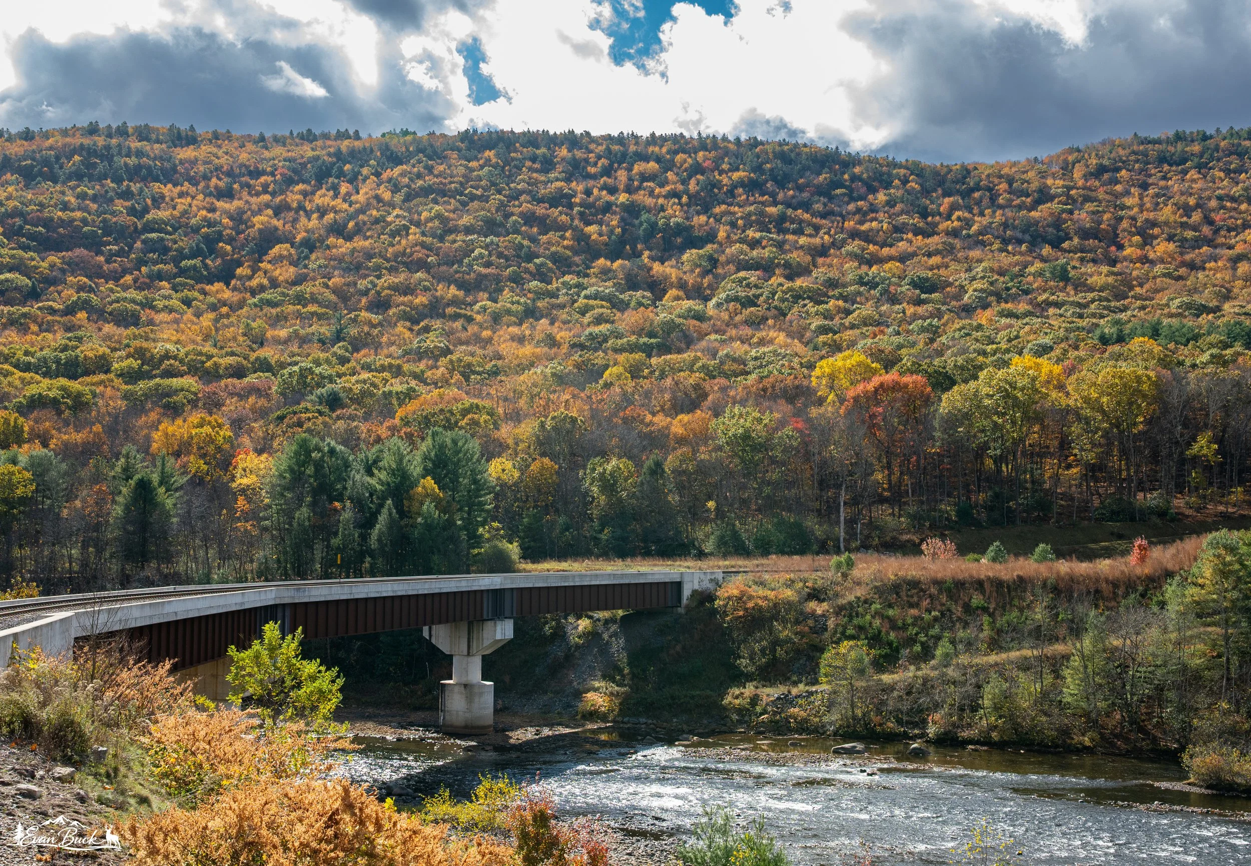 A bridge over a river with a forested hill in autumn behind it, under partly cloudy sky.