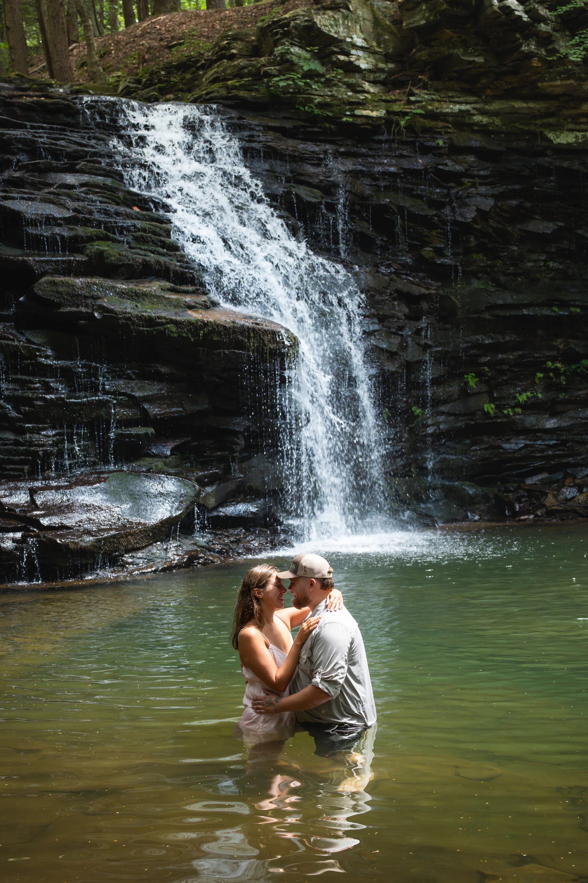 A couple embracing and smiling in a shallow river at the base of a waterfall in a forest.