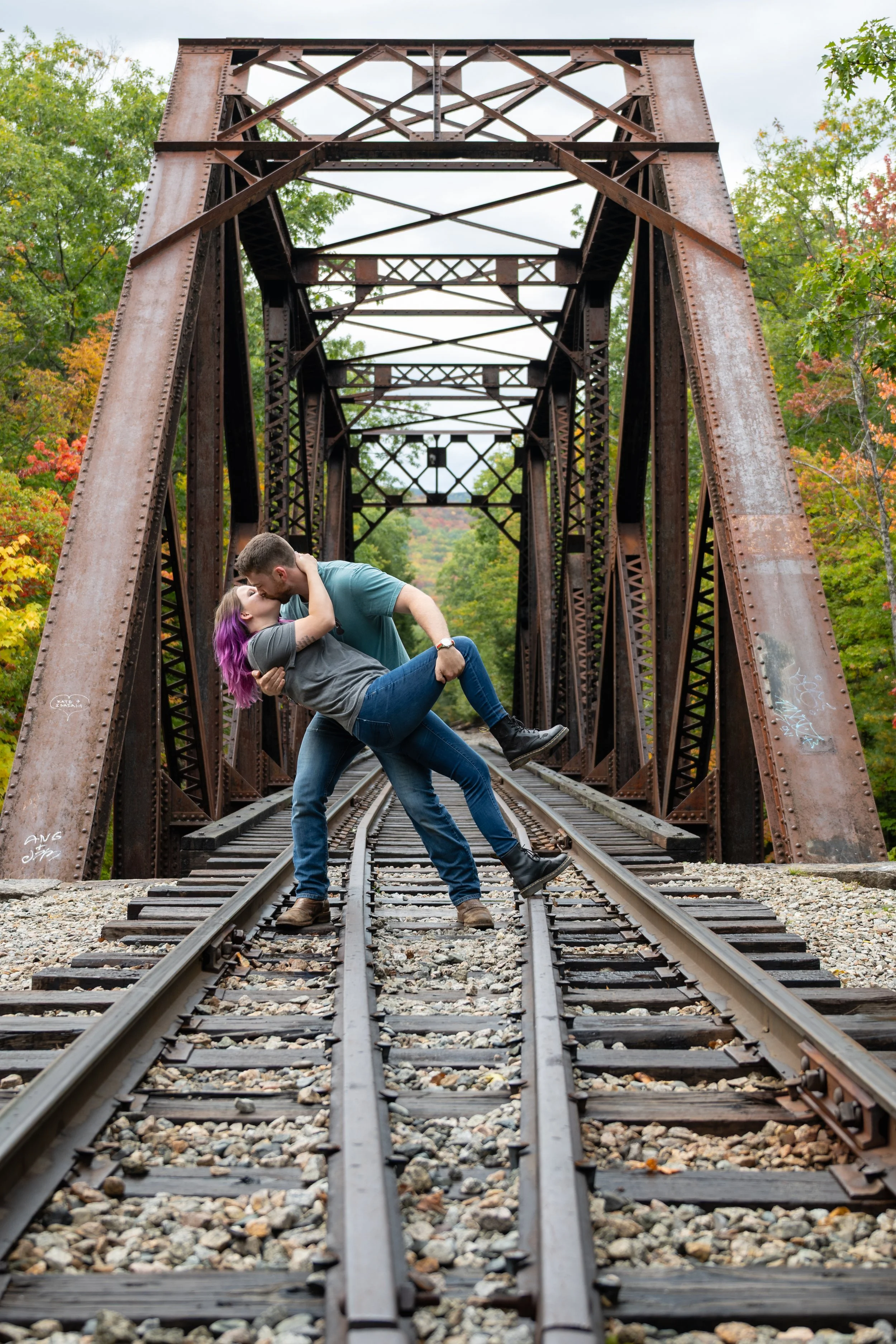 A couple dancing on train tracks under an old rusted metal bridge surrounded by trees with fall foliage.