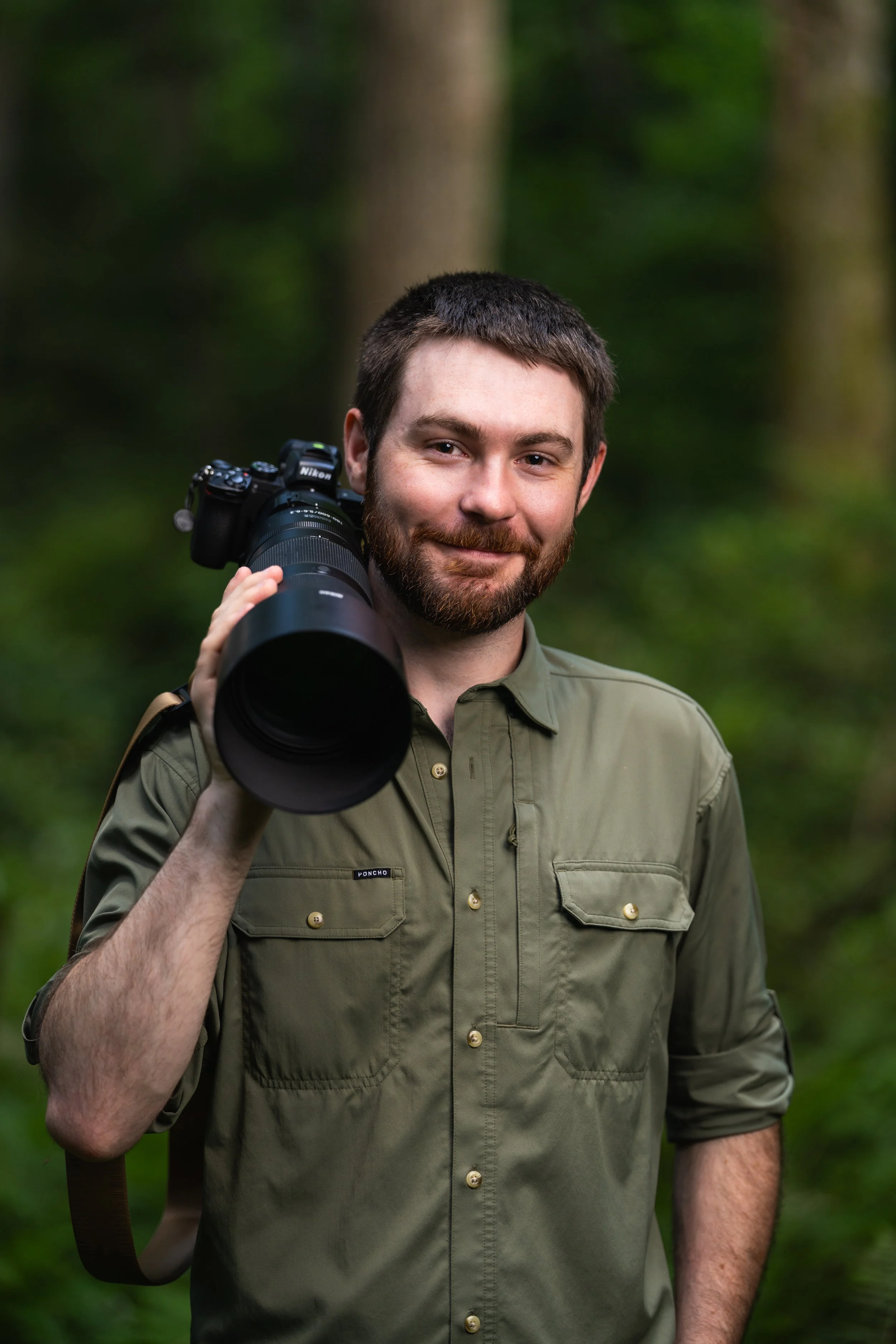 Evan Buck of Evan Buck Photography smiling, holding a camera with a large telephoto lens over his shoulder, outdoors in a forest