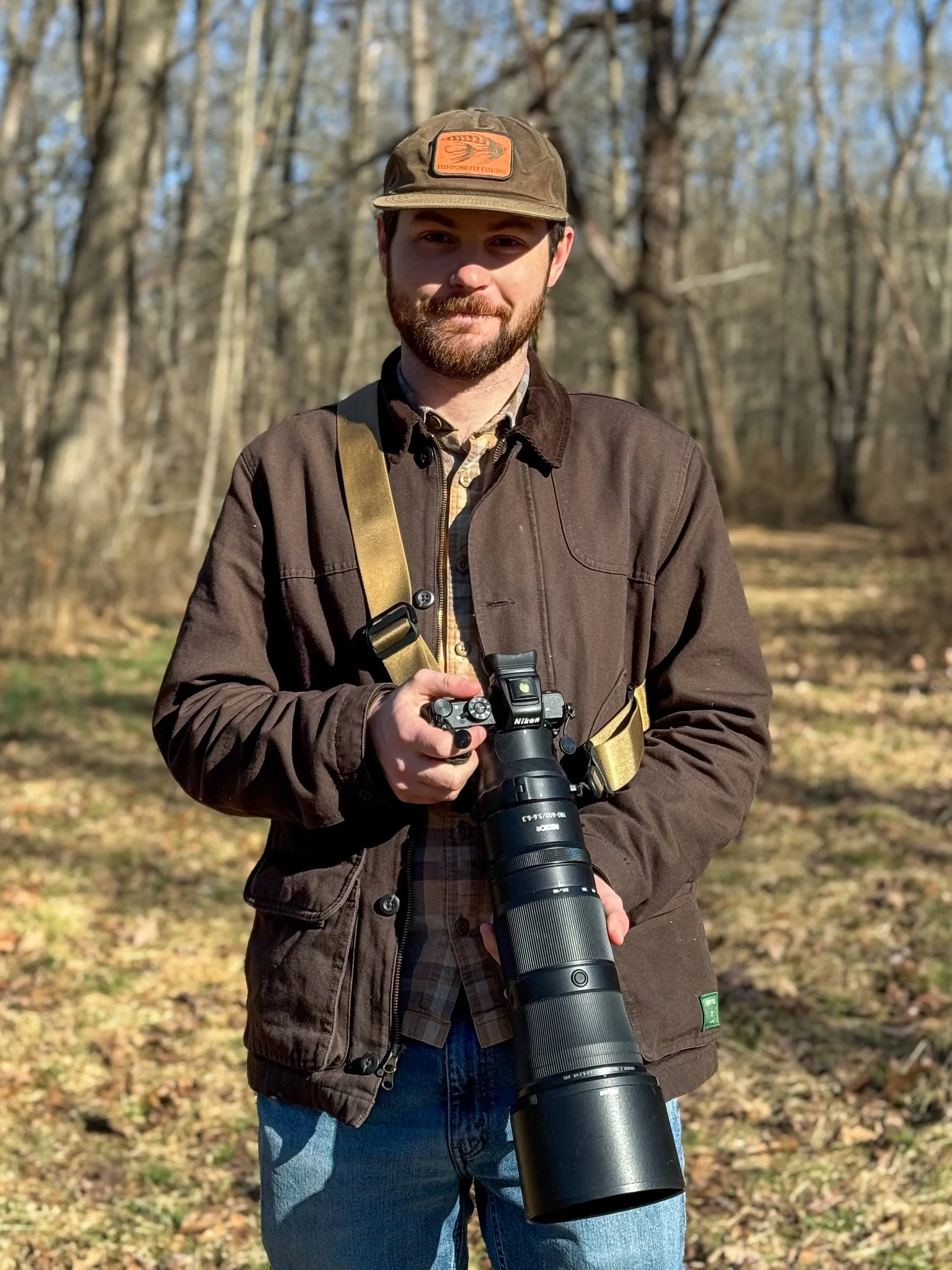 A man standing outdoors in a wooded area, holding a camera with a large telephoto lens, wearing a brown jacket, a plaid shirt, a hat, and jeans.
