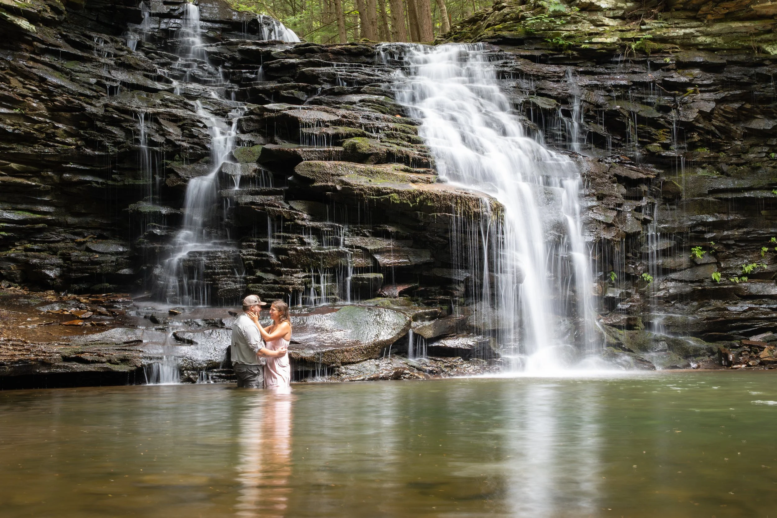 A couple in wedding attire standing in a small lake at the base of a waterfall in a forest.