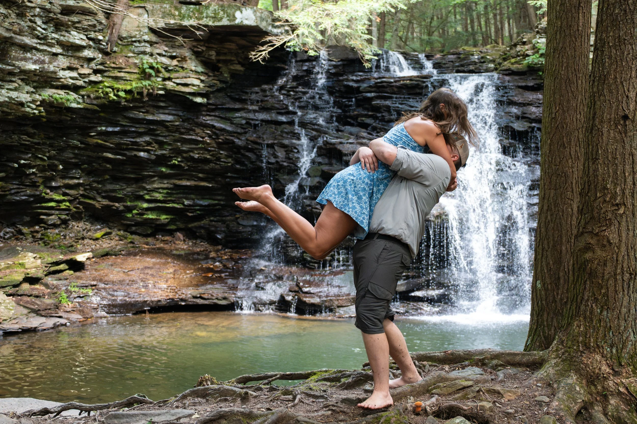 A man lifting a woman in a kiss in front of a small waterfall in a forest, with trees and rocky ground.