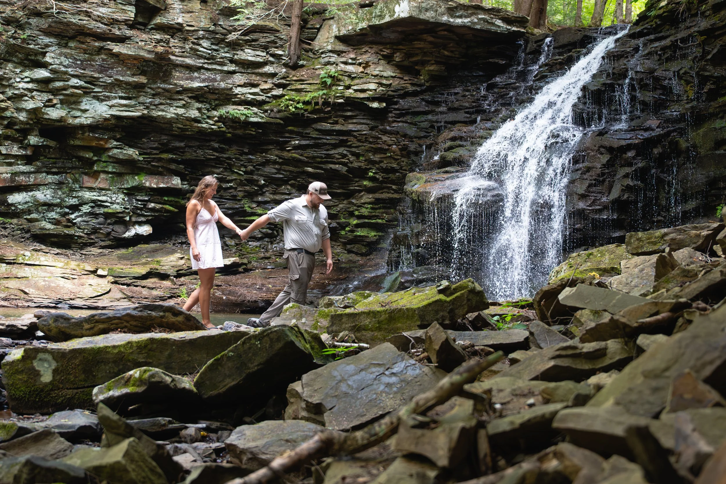A couple holding hands and walking on rocks near a waterfall in a forested area.
