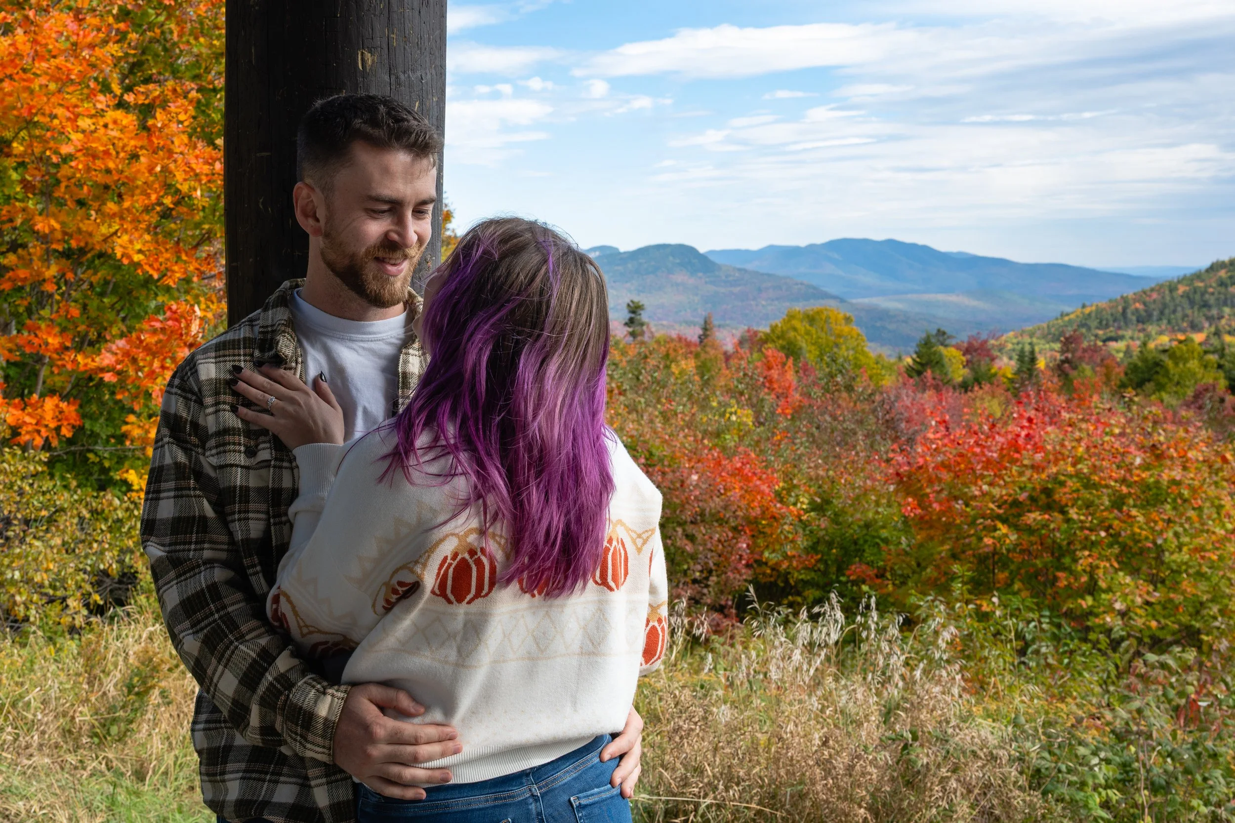 A couple sharing an intimate moment outdoors during autumn. The man has a beard, short hair, and is wearing a plaid shirt over a white T-shirt. The woman has long hair dyed purple, wearing a white sweater with a colorful pattern. They are standing ne