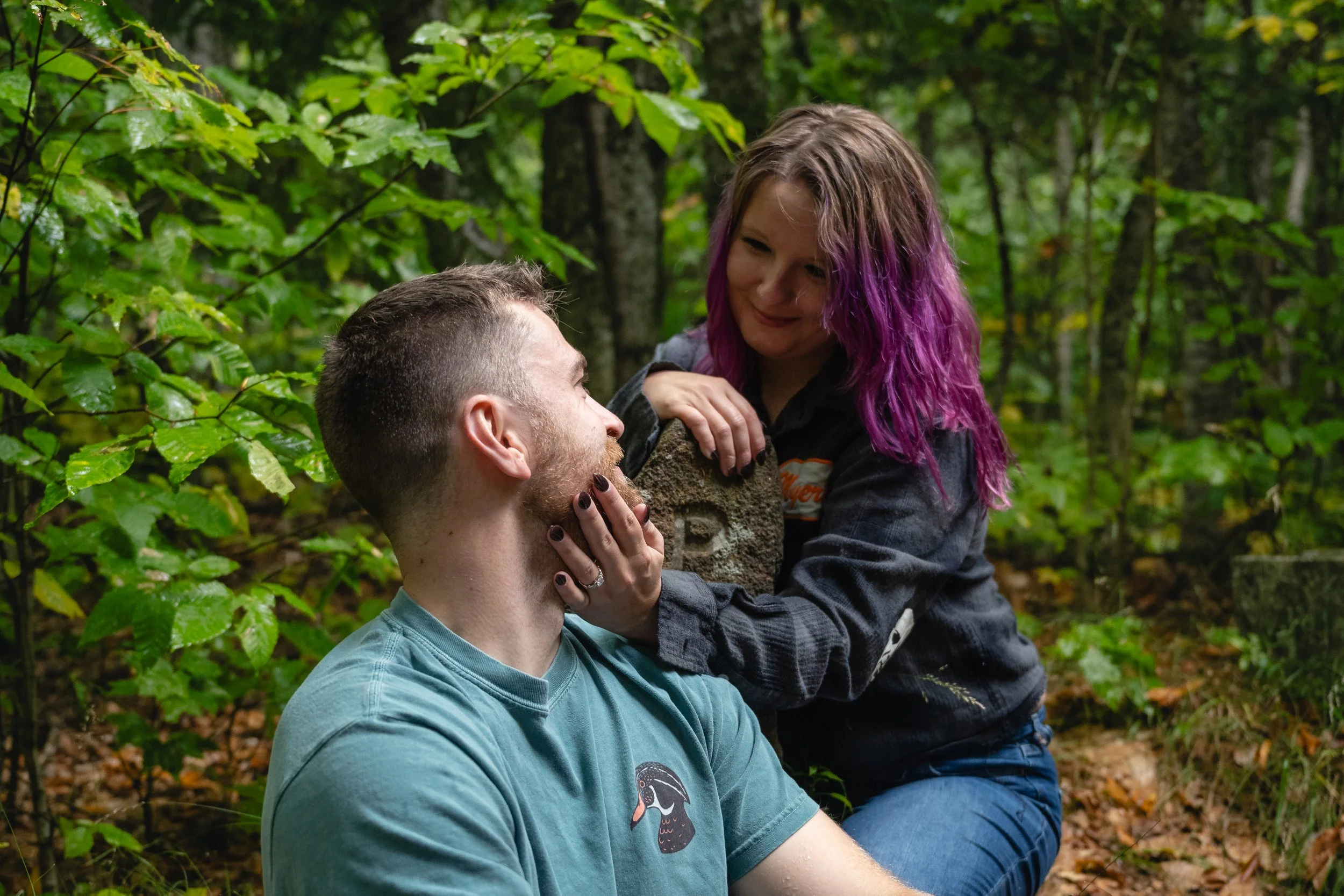 A man with short hair and a beard sitting outdoors in a forest, looking up at a young woman with purple hair, who is smiling at him with her hand on his face.