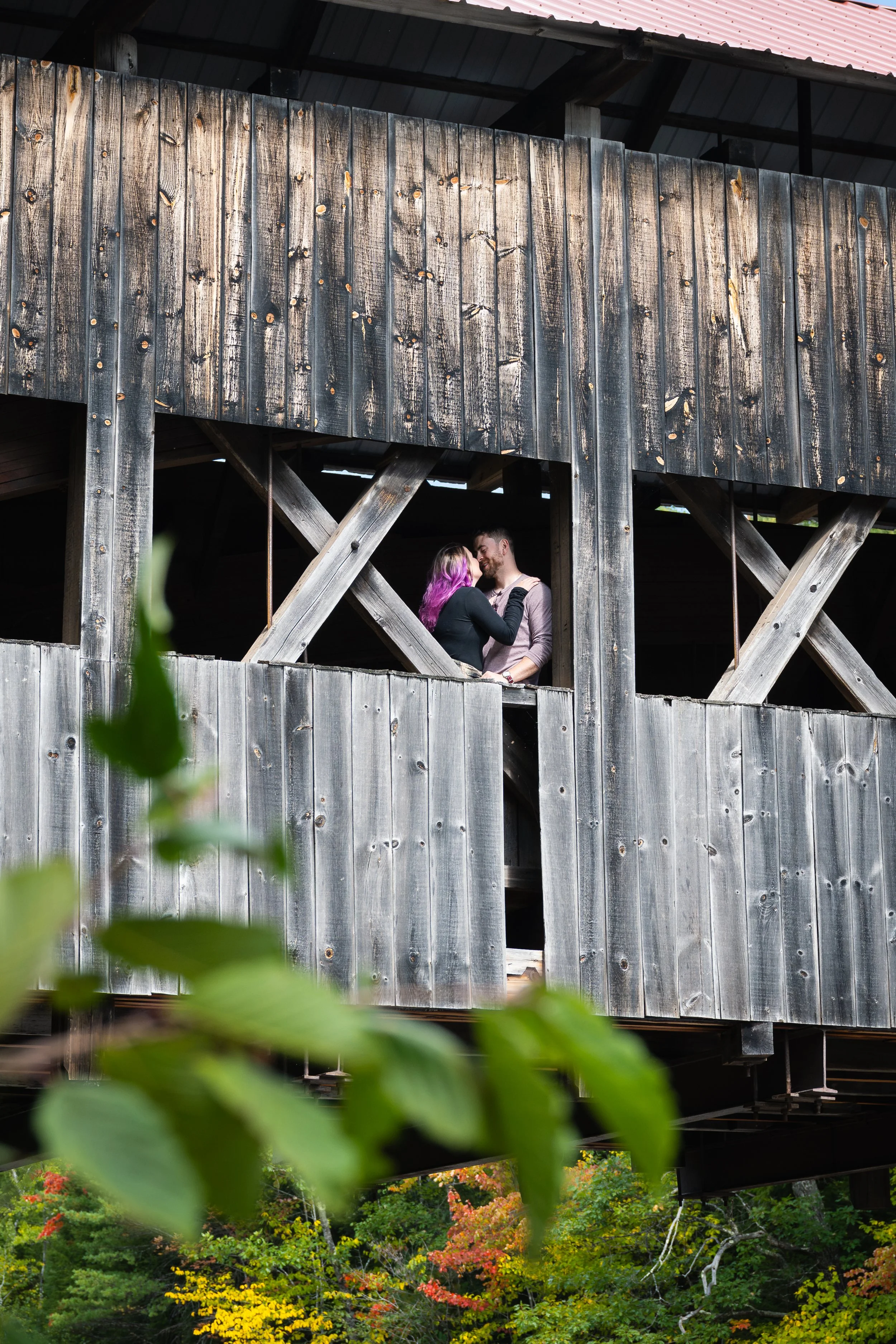 A couple standing and close together framed by a window of an old wooden covered bridge, surrounded by a forest with colorful autumn trees.