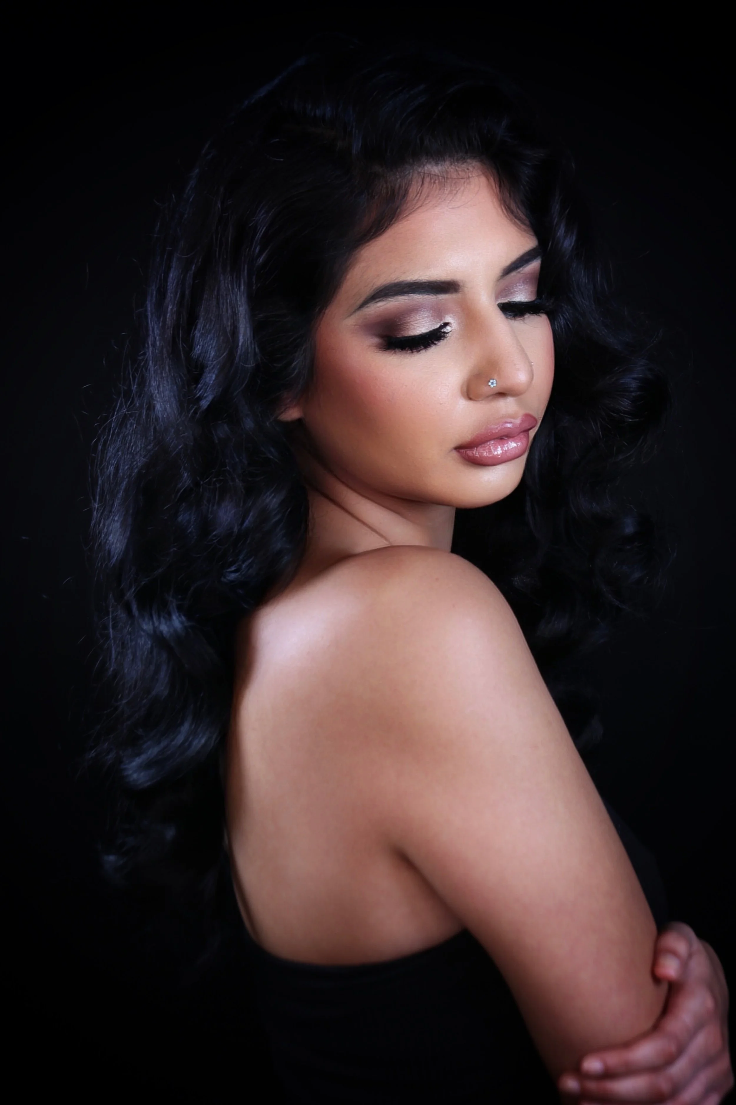 A woman with long black curly hair and makeup looking down with eyes closed, with a small nose piercing, against a black background.