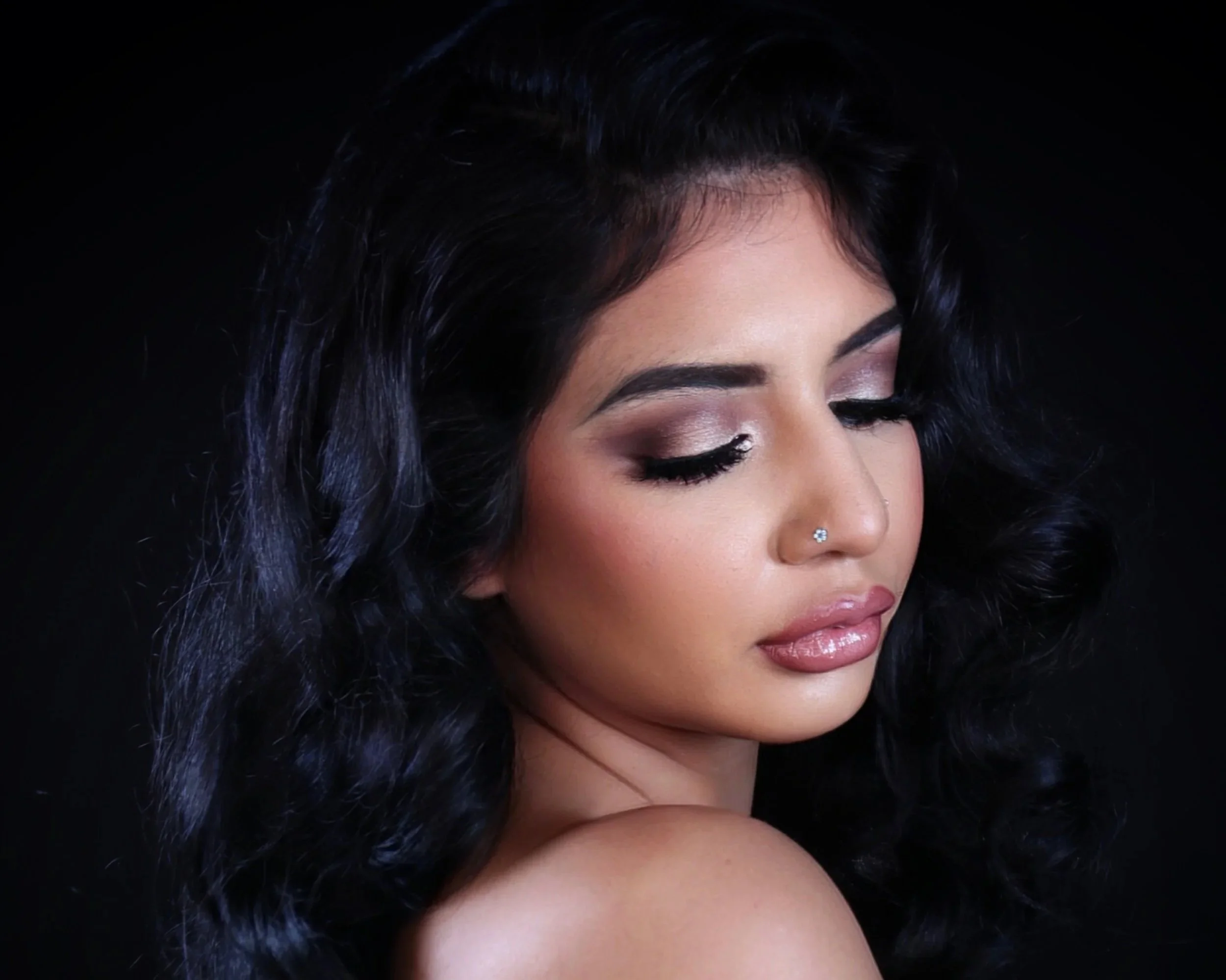 Close-up of a woman with dark, wavy hair, makeup, and a nose piercing, with her eyes closed against a black background.