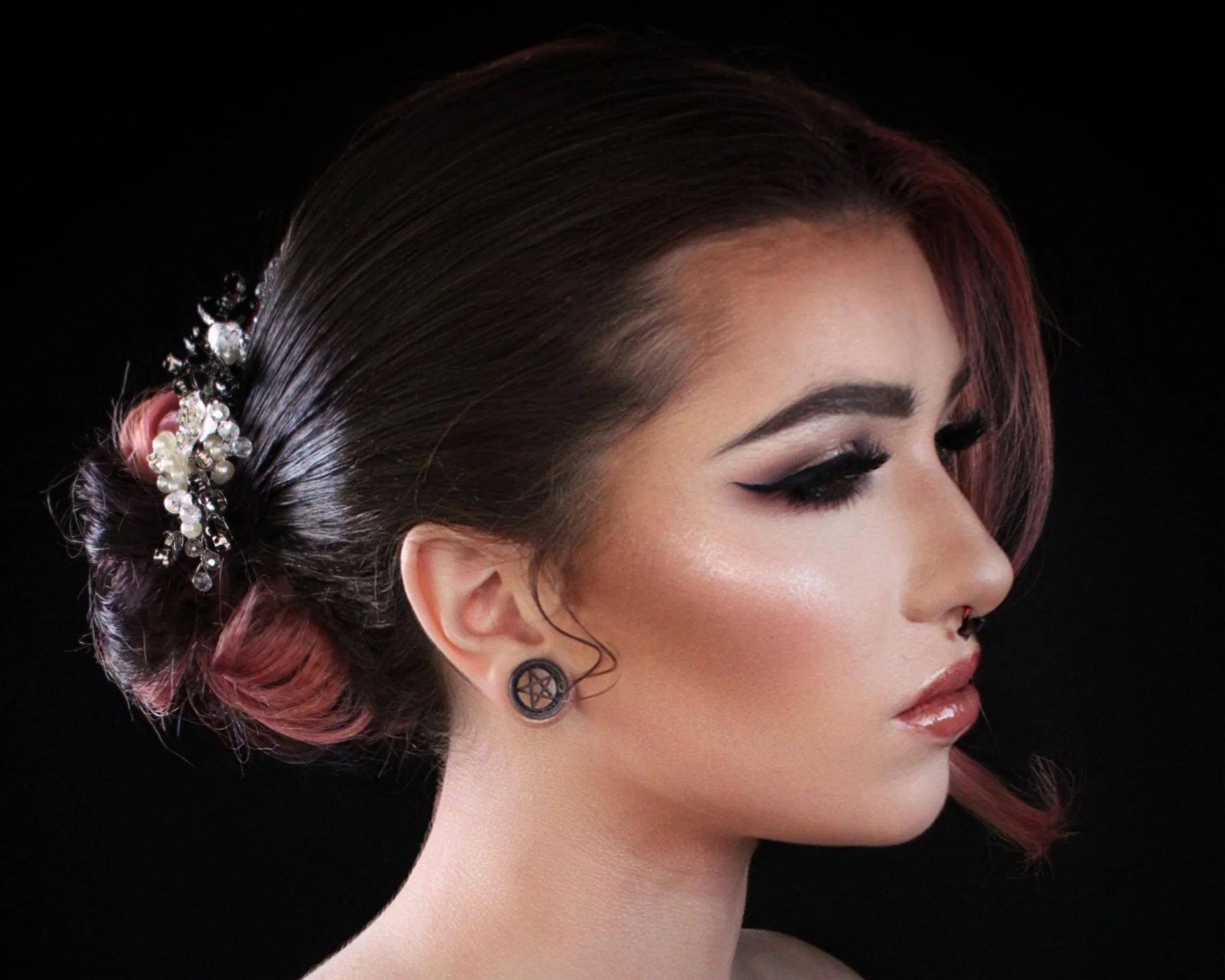 Side profile of a woman with styled dark and pink hair, wearing pentagram earrings and decorated hair accessory, against a black background.