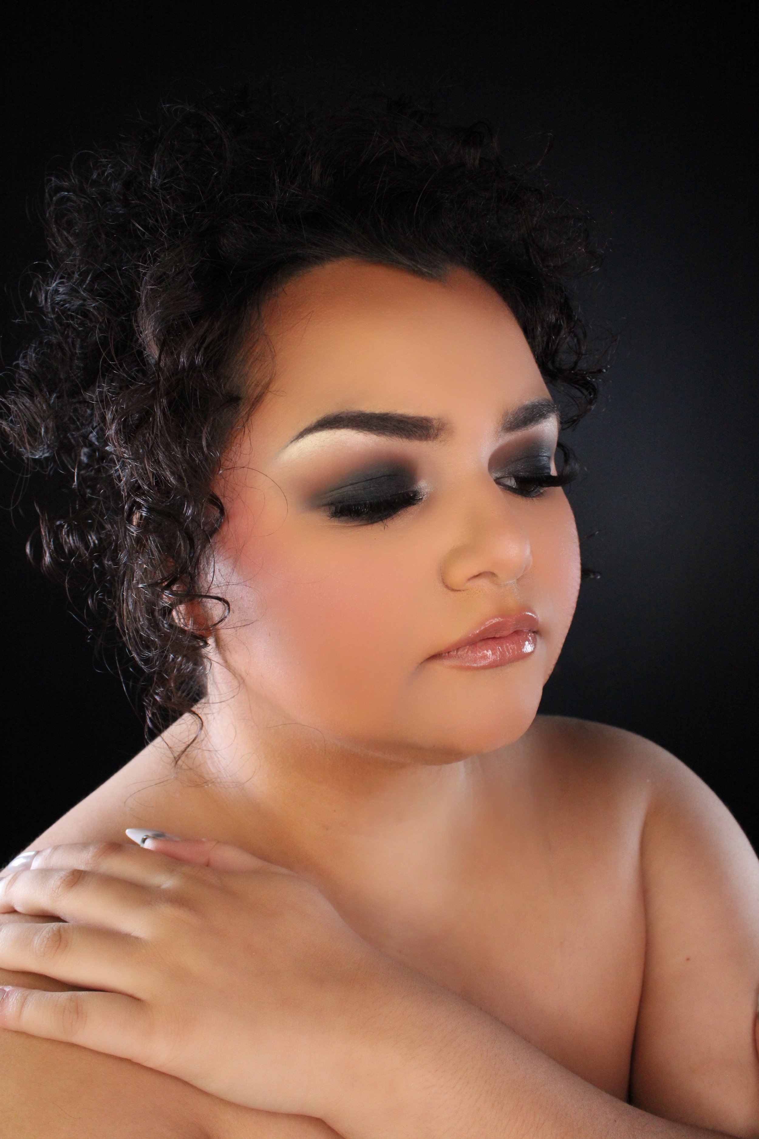 Close-up portrait of a woman with curly hair, wearing makeup, with eyes closed and one hand resting on her shoulder.