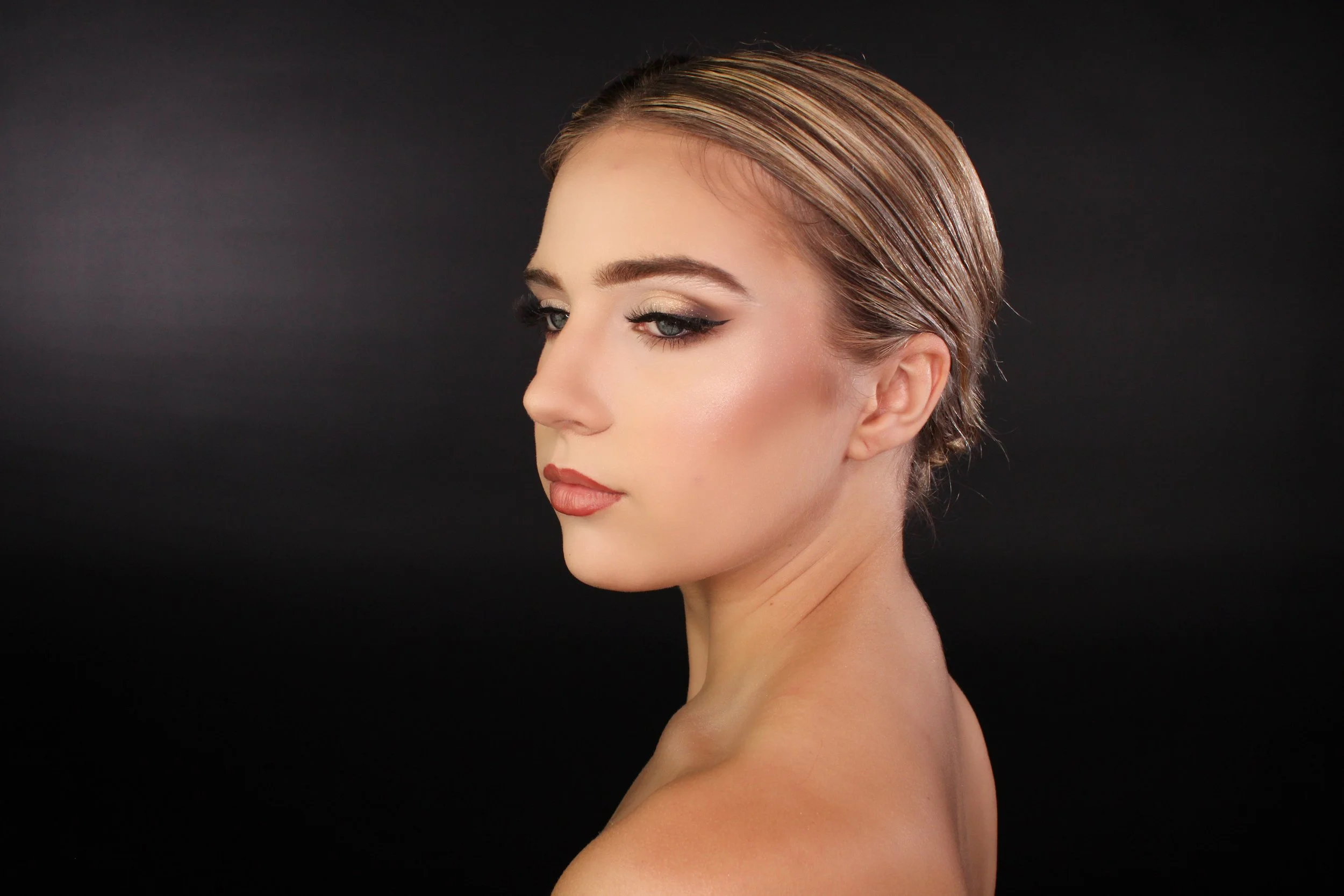 Profile of a woman with makeup and slicked-back hair against a black background.