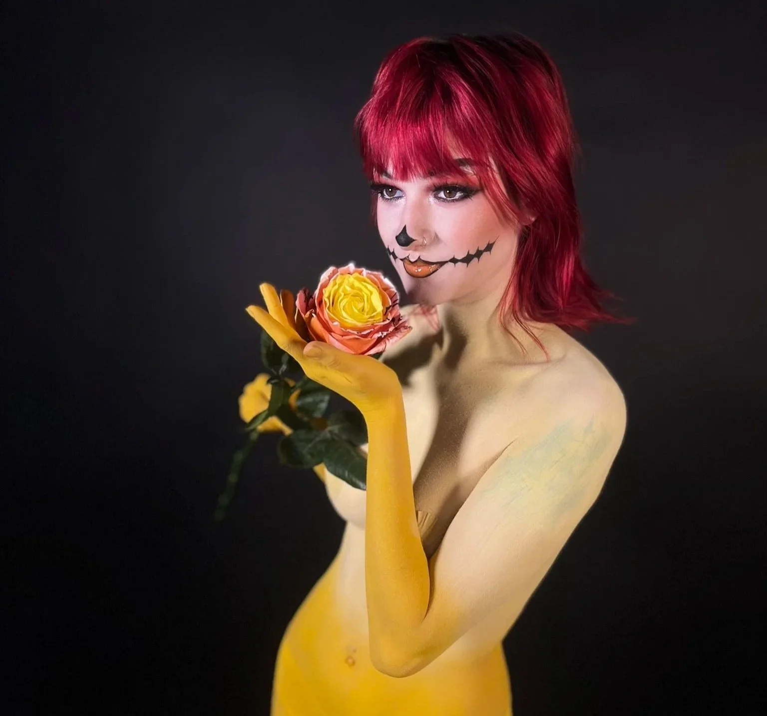 A person with red hair and face paint resembling a skeleton smile is holding a pink and yellow rose, with a black background.
