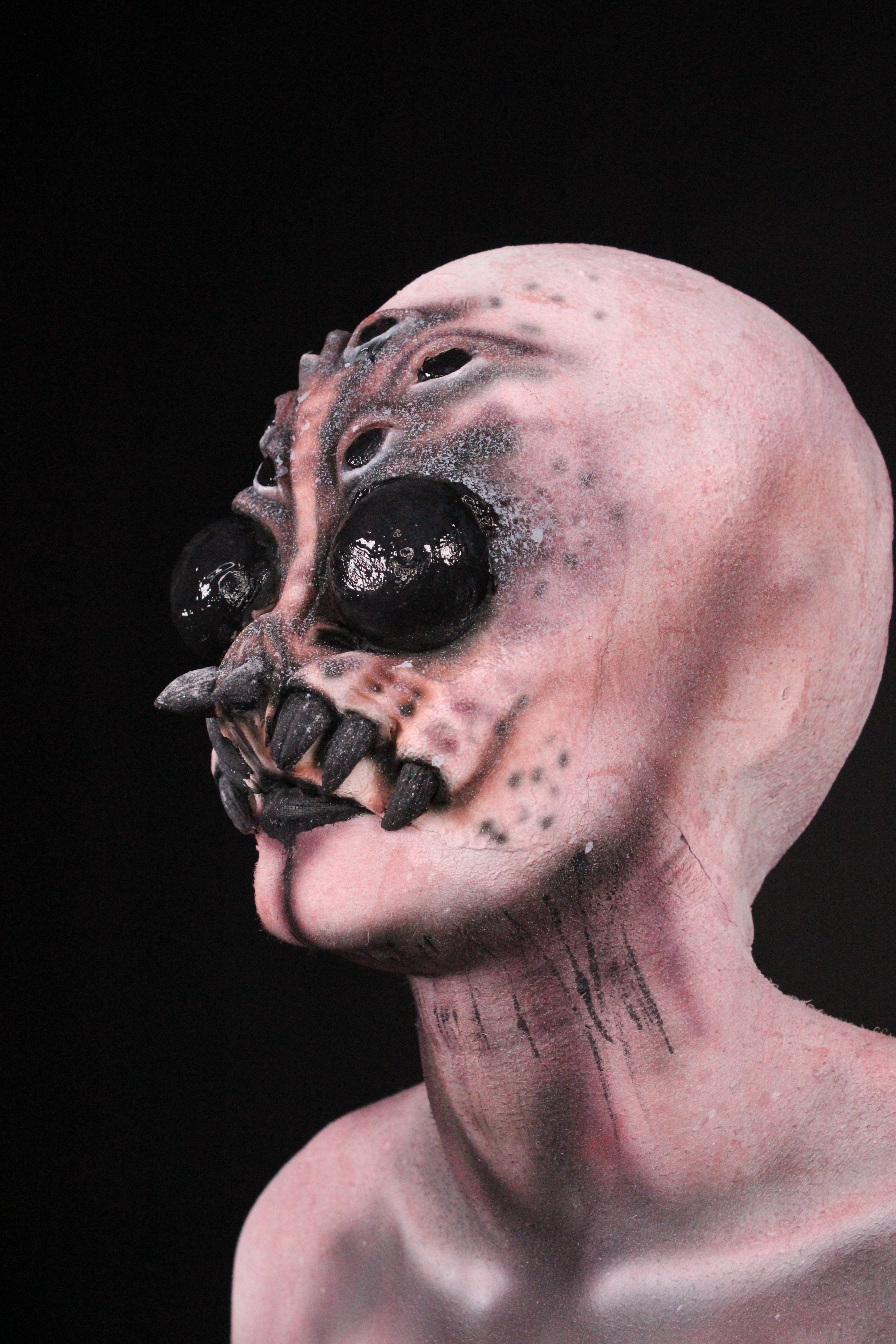 A surreal sculpture combining a human head with a skull that has a third eye on the forehead, black eye sockets, and black teeth, against a dark background.