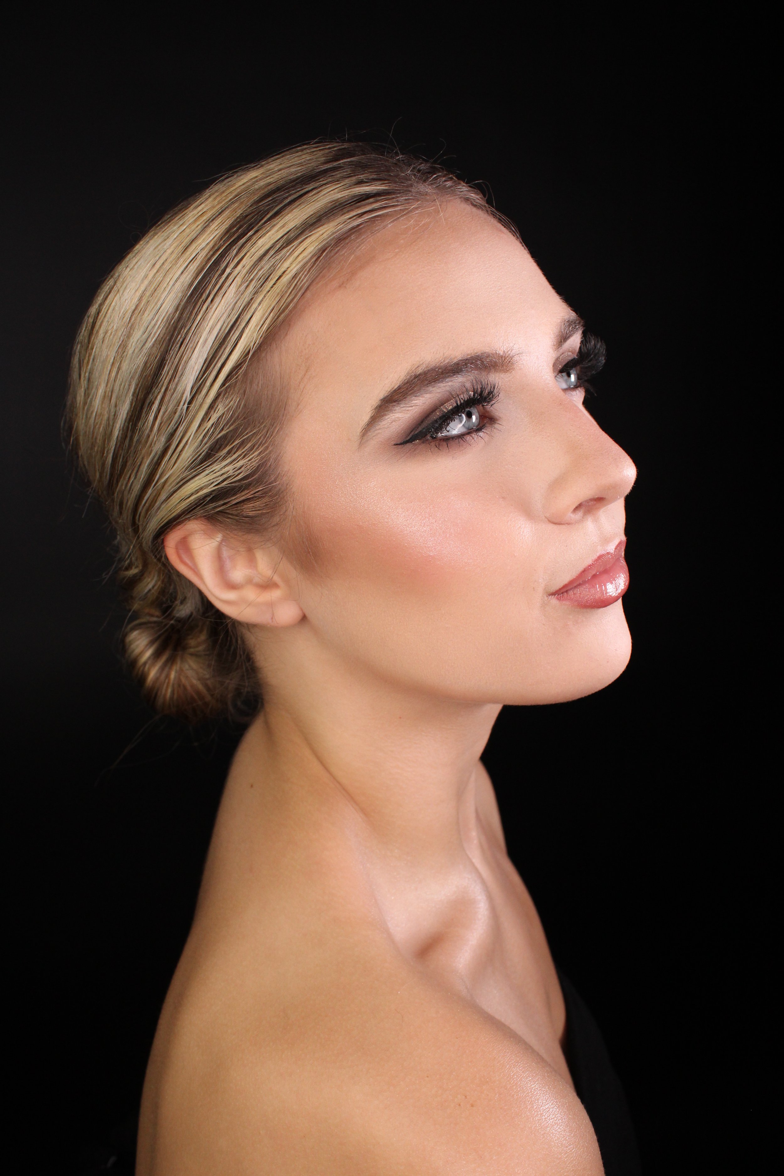 A woman with blonde hair styled in a side bun, wearing makeup with dark eyeliner and nude lipstick, looking upwards against a black background.