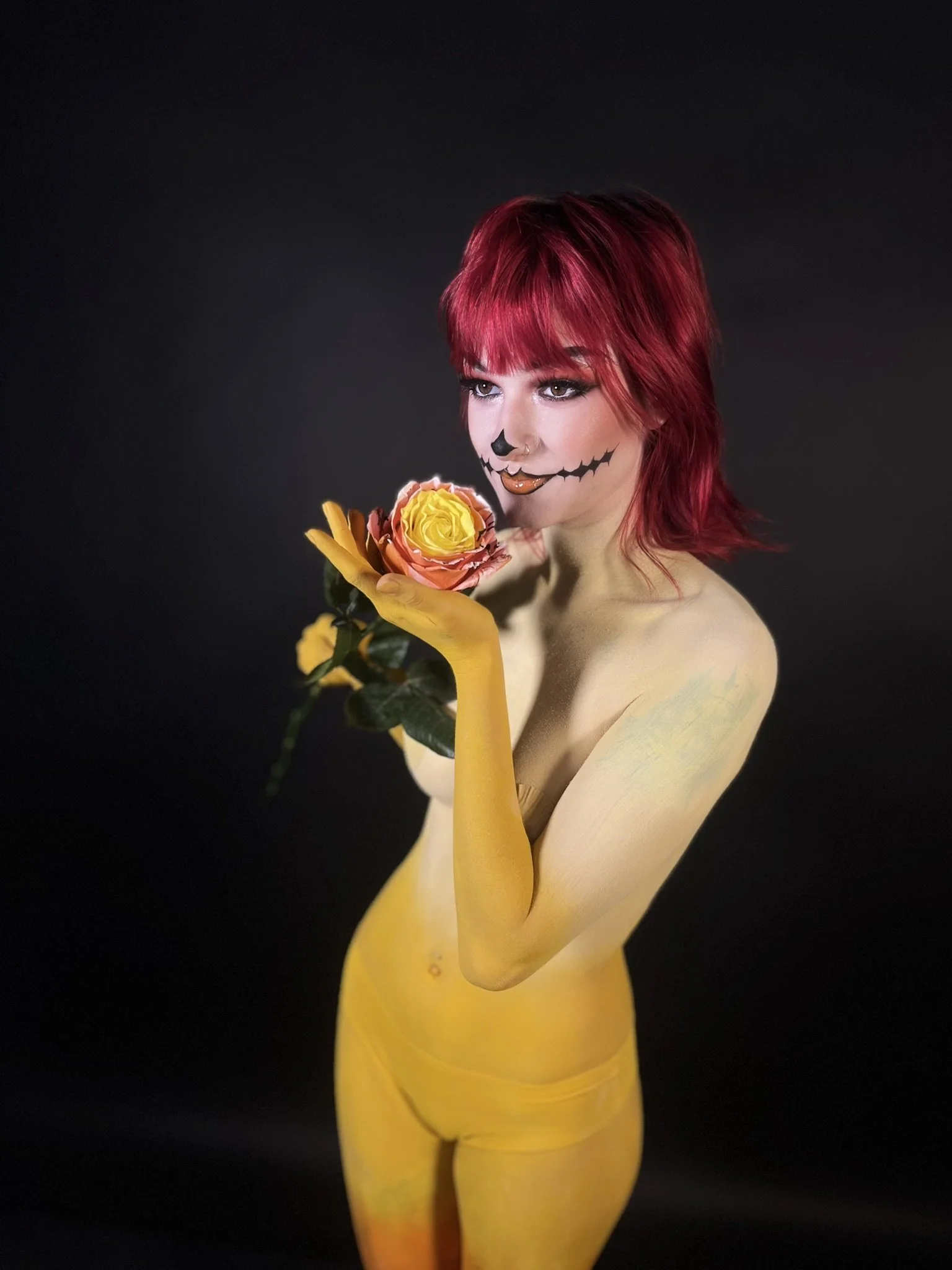 A woman with red hair painted with Halloween makeup, holding a pink and yellow rose, standing against a black background.