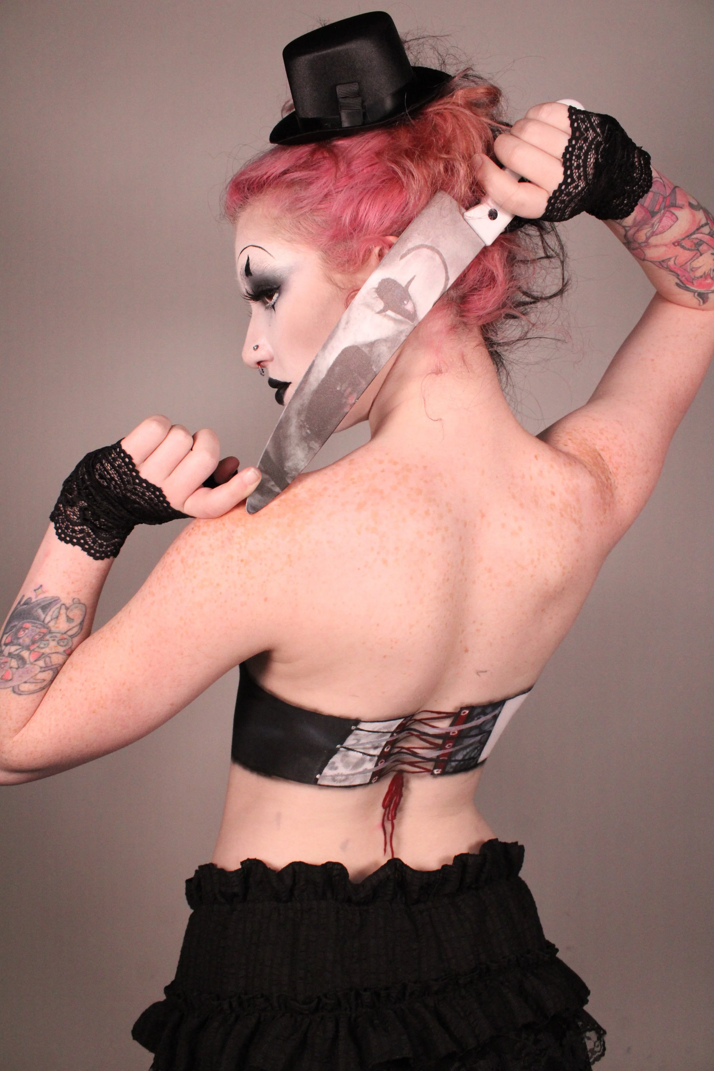 A woman with pink and black hair, wearing a small black hat, displays tattoos on her arms and back, holds a large knifed over her shoulder, and wears black lace gloves and a black corset with a lace-up back.