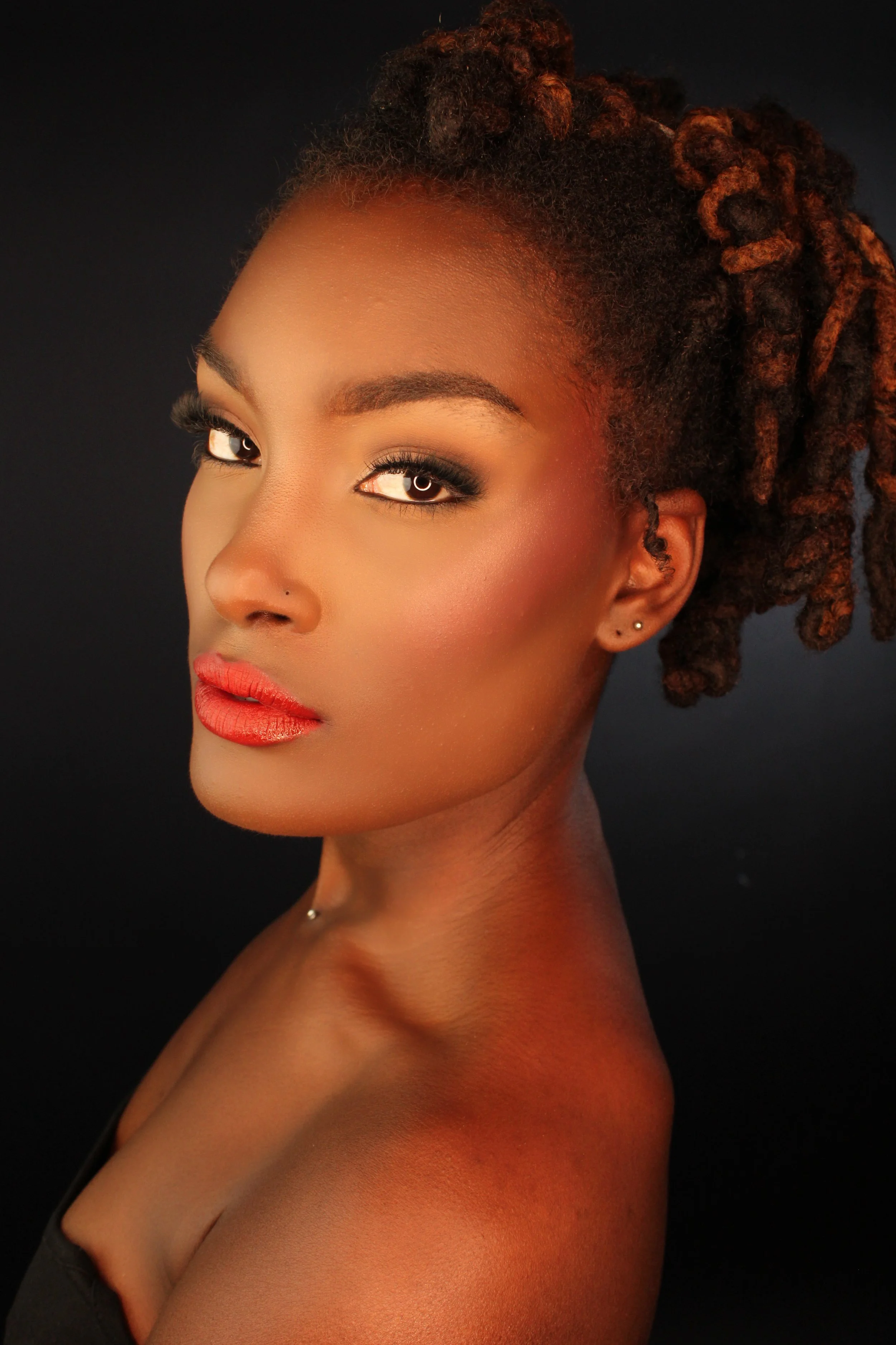 A close-up portrait of a woman with styled dreadlocks, radiant makeup, and a confident expression against a dark background.