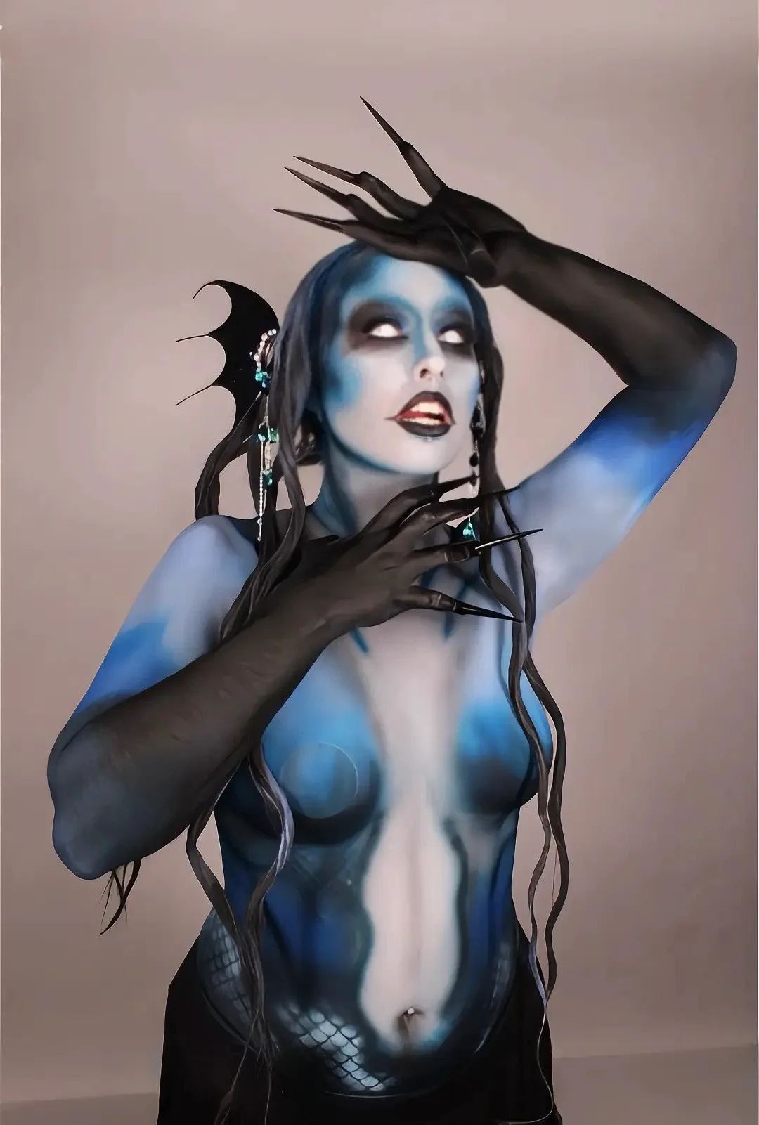 A woman painted in blue and black body paint with long dark hair. She has dark makeup, wearing dangling earrings, with long pointed black nails, and a black wing earring with bead accents. She poses with her hands near her face and head, against a plain background.