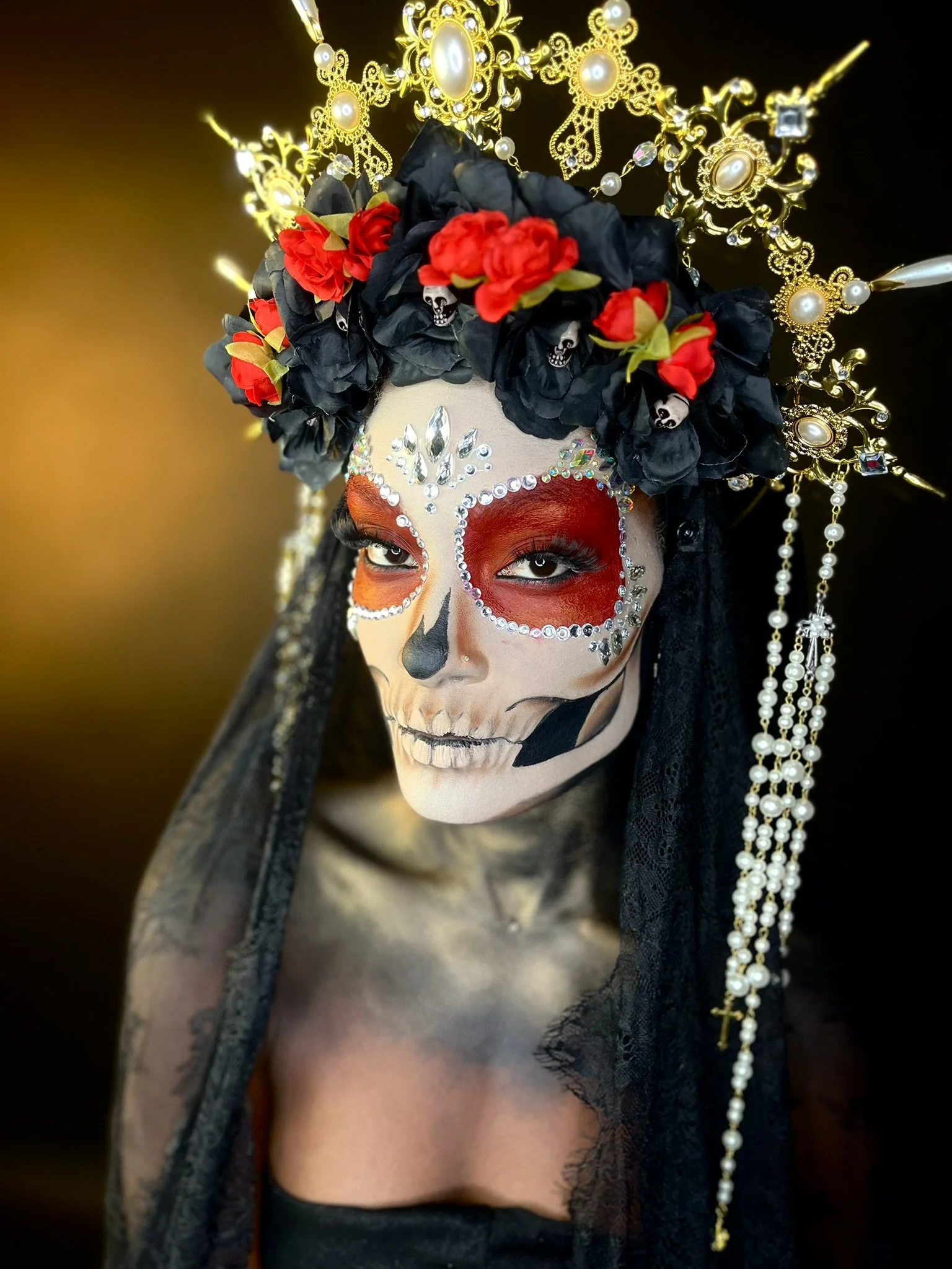 Woman dressed in Day of the Dead costume with skull face paint, red eye circles, floral headpiece, and gold jewelry headpiece, with black lace covering her shoulders.