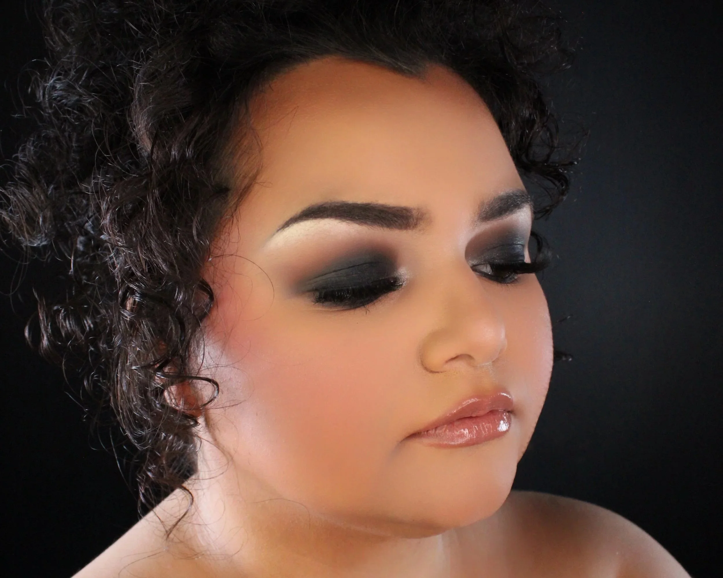 Close-up of a woman's face with makeup, curly dark hair, and closed eyes, against a black background.