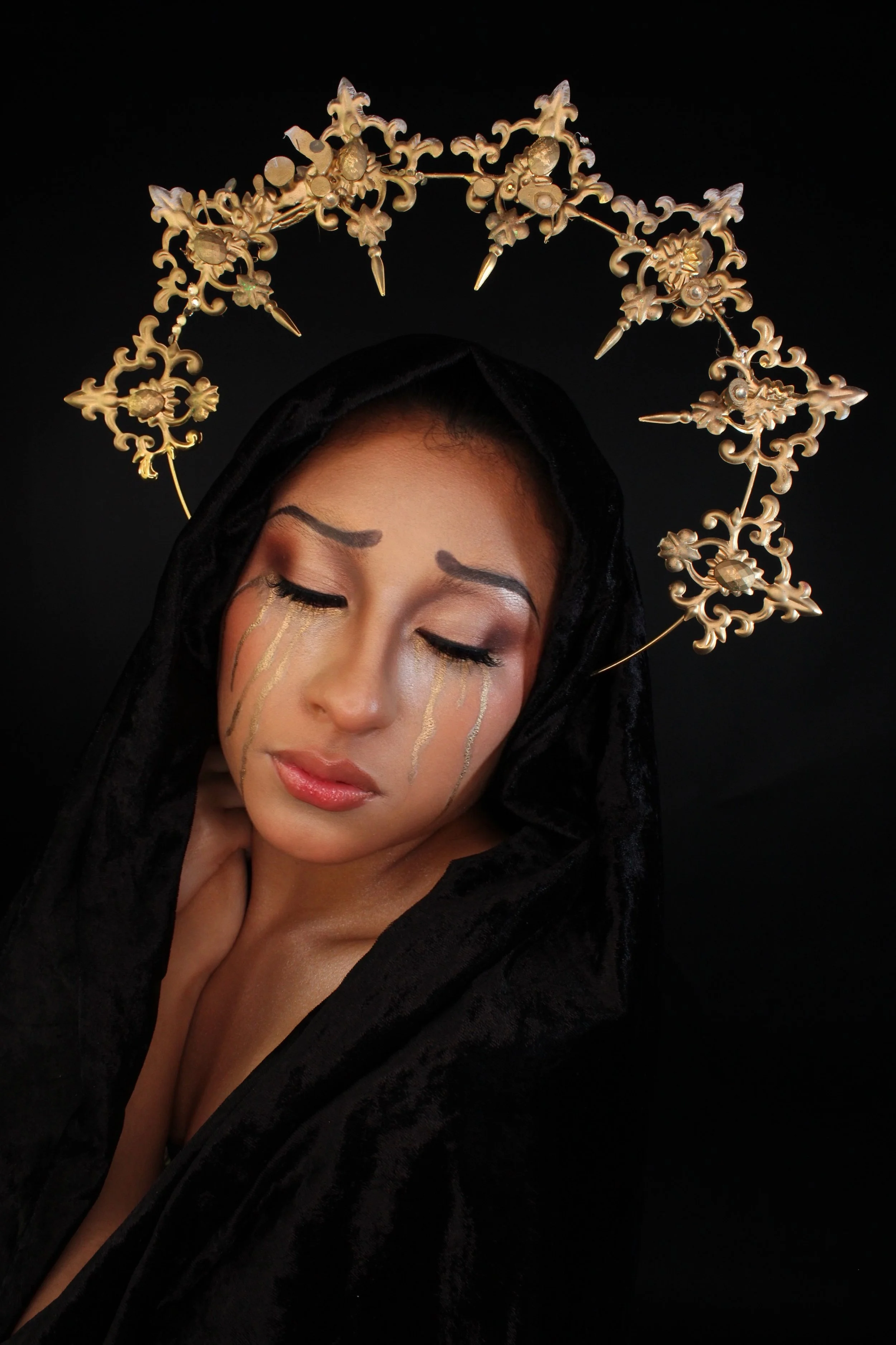 A woman with closed eyes wearing gold jewelry and black clothing against a black background.