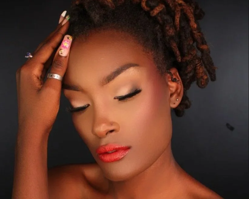 A woman with styled natural hair, closed eyes, and makeup including red lipstick, with her hand resting on her forehead against a dark background.