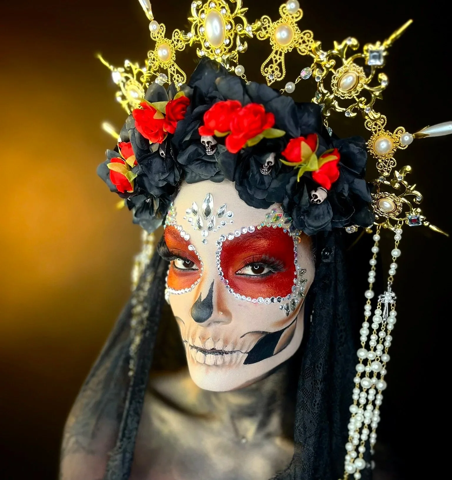 A person with intricate sugar skull makeup, wearing a black and red floral headdress with gold and pearl embellishments, and a black veil.