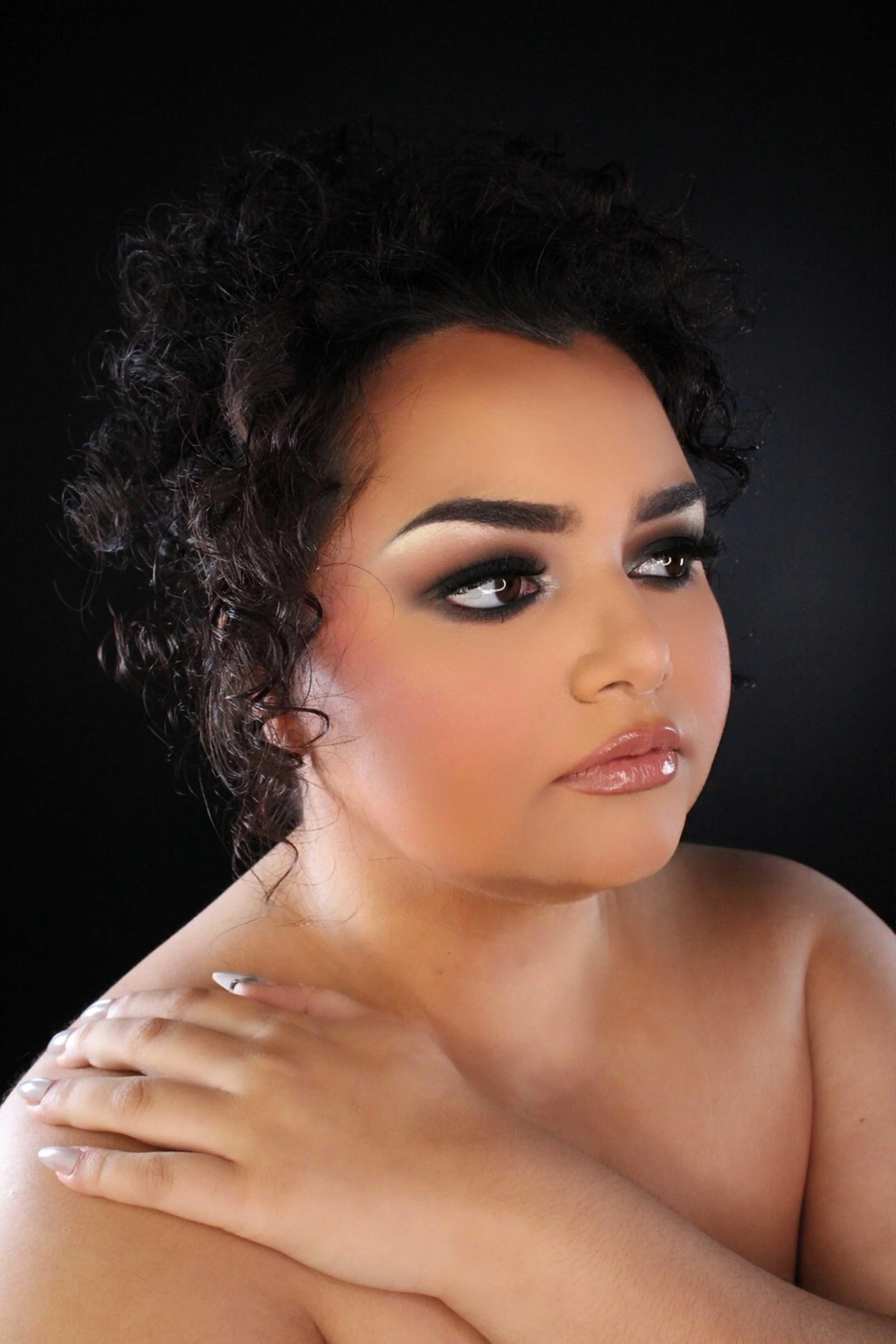 Close-up portrait of a woman with short curly black hair and makeup, resting her arms on her shoulders against a black background.