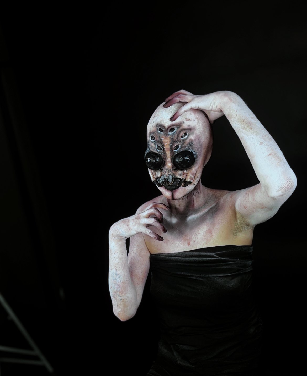 Person in black clothing wearing a detailed creepy mask with multiple eyes and black prominent features, posing with hand on head against a black background.