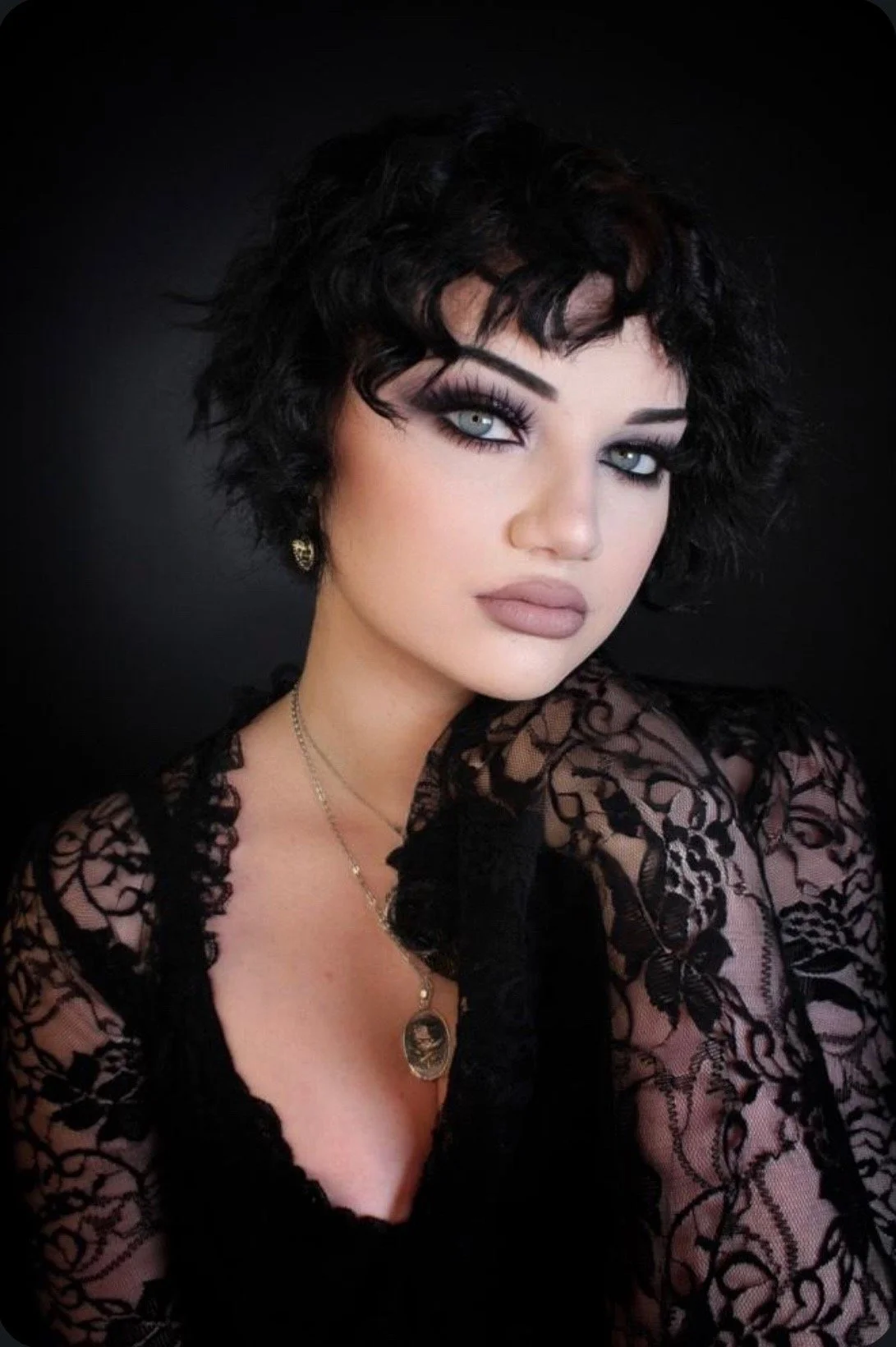 Portrait of a woman with short curly black hair, gray eyes, and makeup, wearing lace clothing and jewelry against a black background.