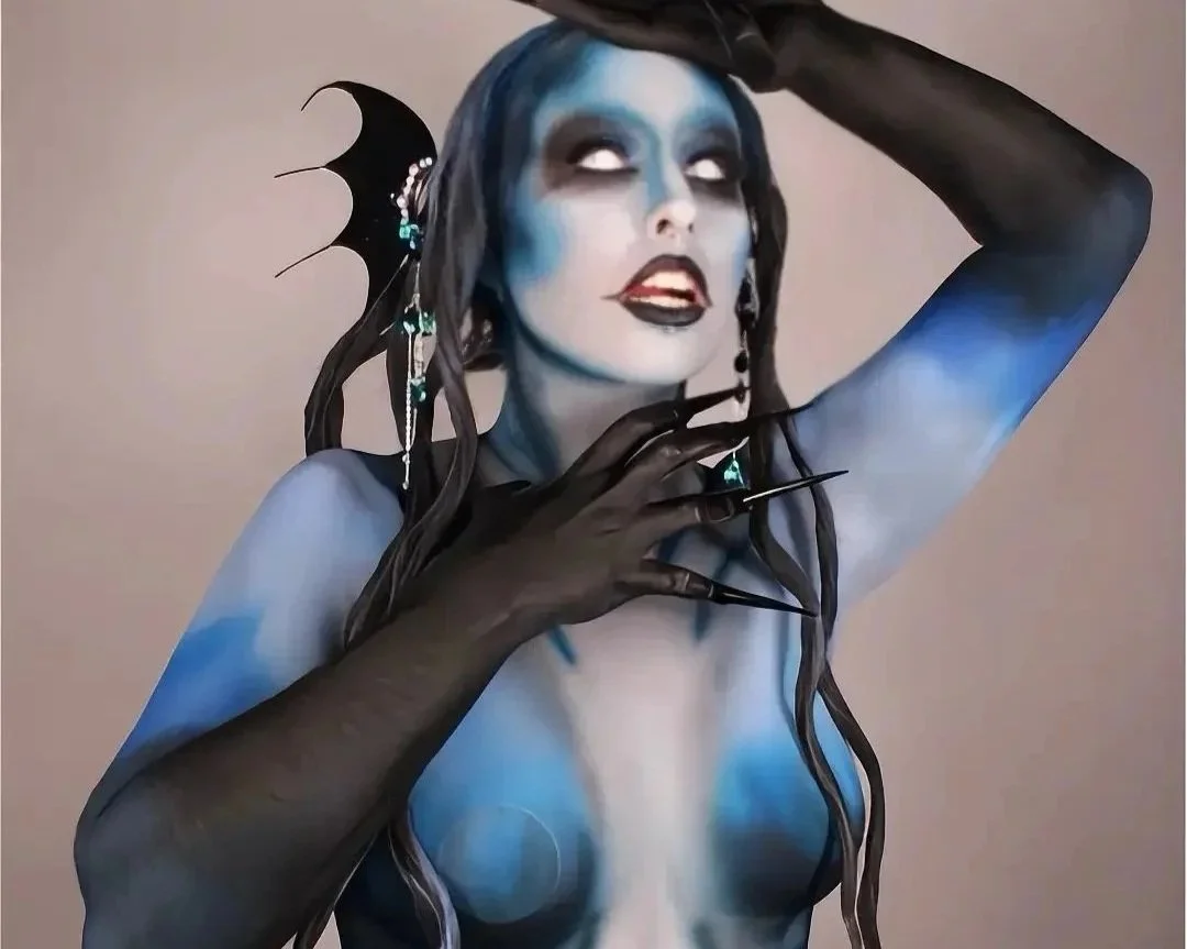 A person painted blue with black accents, wearing dramatic makeup, long black gloves, and black pointed nails. They have long dark hair adorned with a black bat-shaped accessory and dangling earrings, striking a pose with one hand touching their face and the other behind their head.