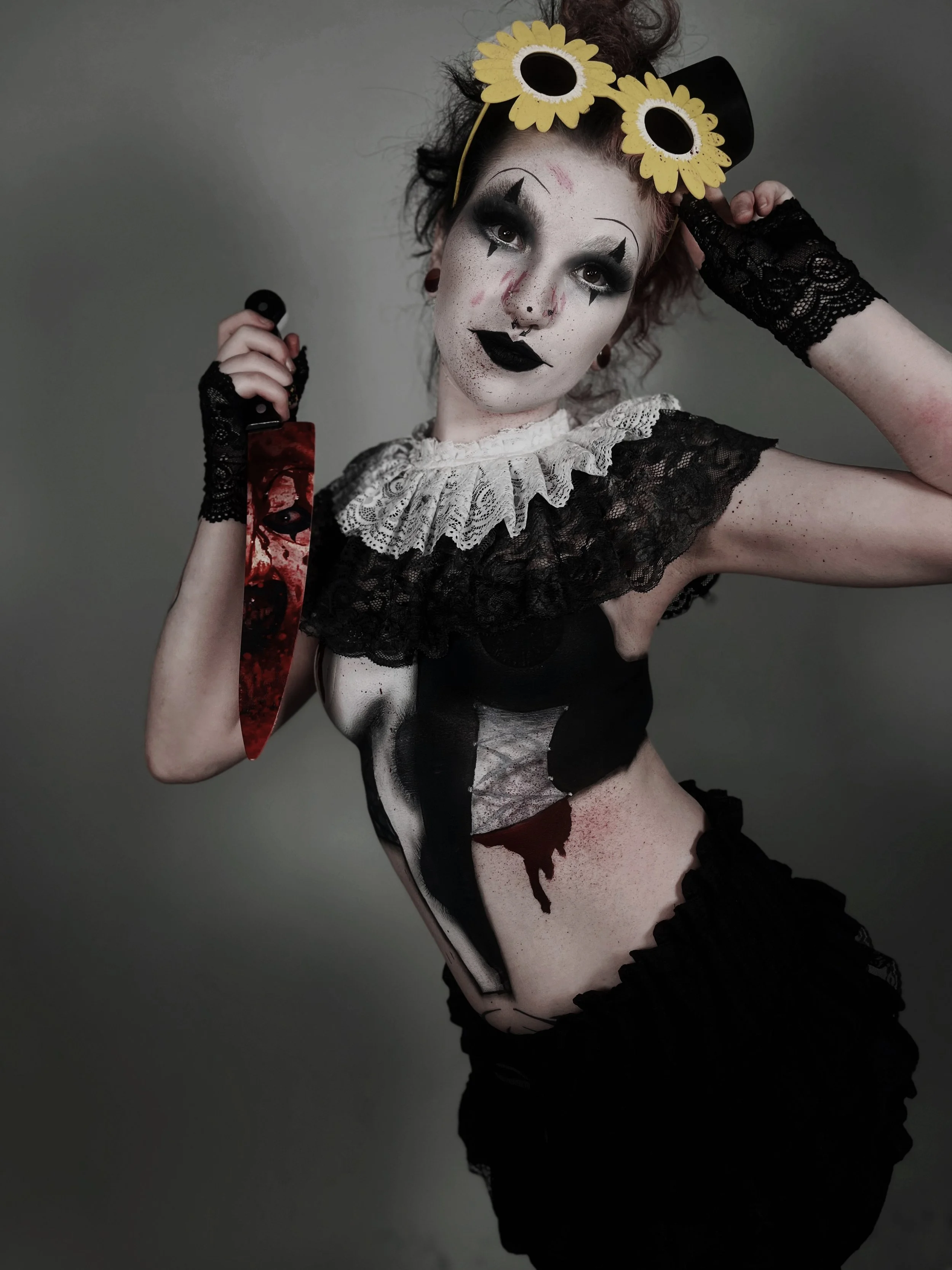 Person dressed as a creepy clown with clown makeup, a sunflower headband, a black dress with lace, holding a bloody knife, against a plain background.