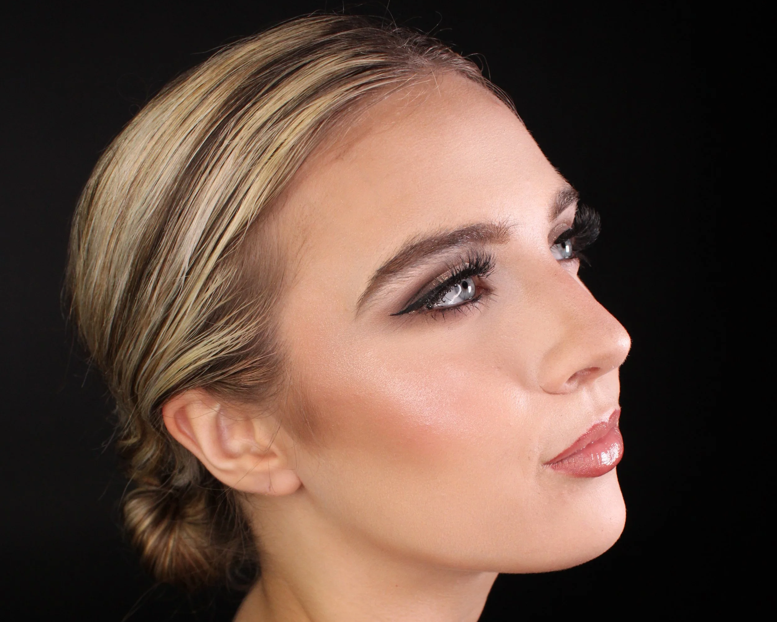 A close-up profile of a woman with blonde hair styled in an elegant updo, wearing makeup with emphasis on dark eyeliner, long lashes, and glossy lips, against a black background.