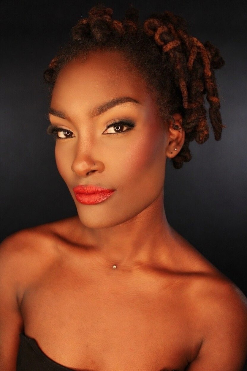 A woman with dreadlocks styled to one side, wearing makeup with bold red lipstick, dark eye makeup, small earrings, and a tiny piercing on her collarbone, against a dark background.