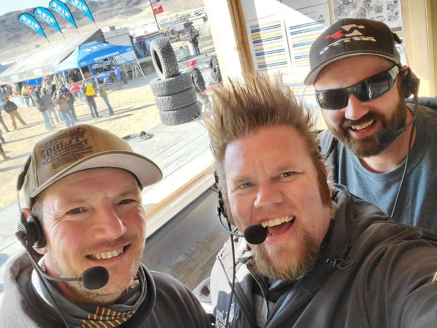 Three smiling men wearing headsets with microphones taking a selfie inside a race station. Outside, a sandy area with tires stacked and people, race flags, and canopies in the background.
