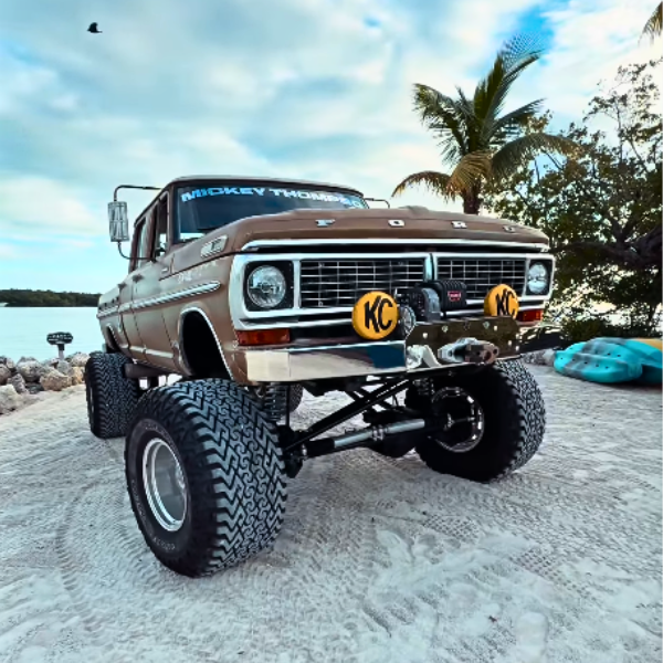 A vintage Ford truck with large off-road tires, parked on a sandy beach with palm trees and a cloudy sky in the background.