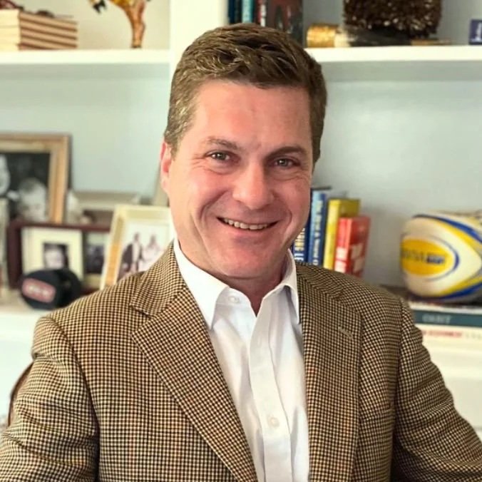 A smiling man with short brown hair wearing a brown checked blazer and white shirt, sitting in a home office with shelves of books, photos, and a rugby ball in the background.