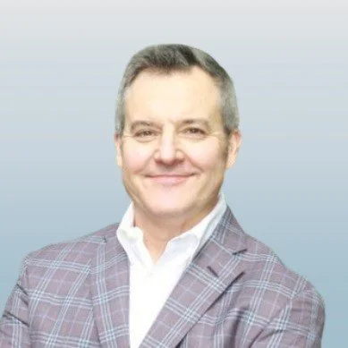 A professional man with short gray hair, wearing a checkered blazer and white collared shirt, smiling against a gradient light blue background.