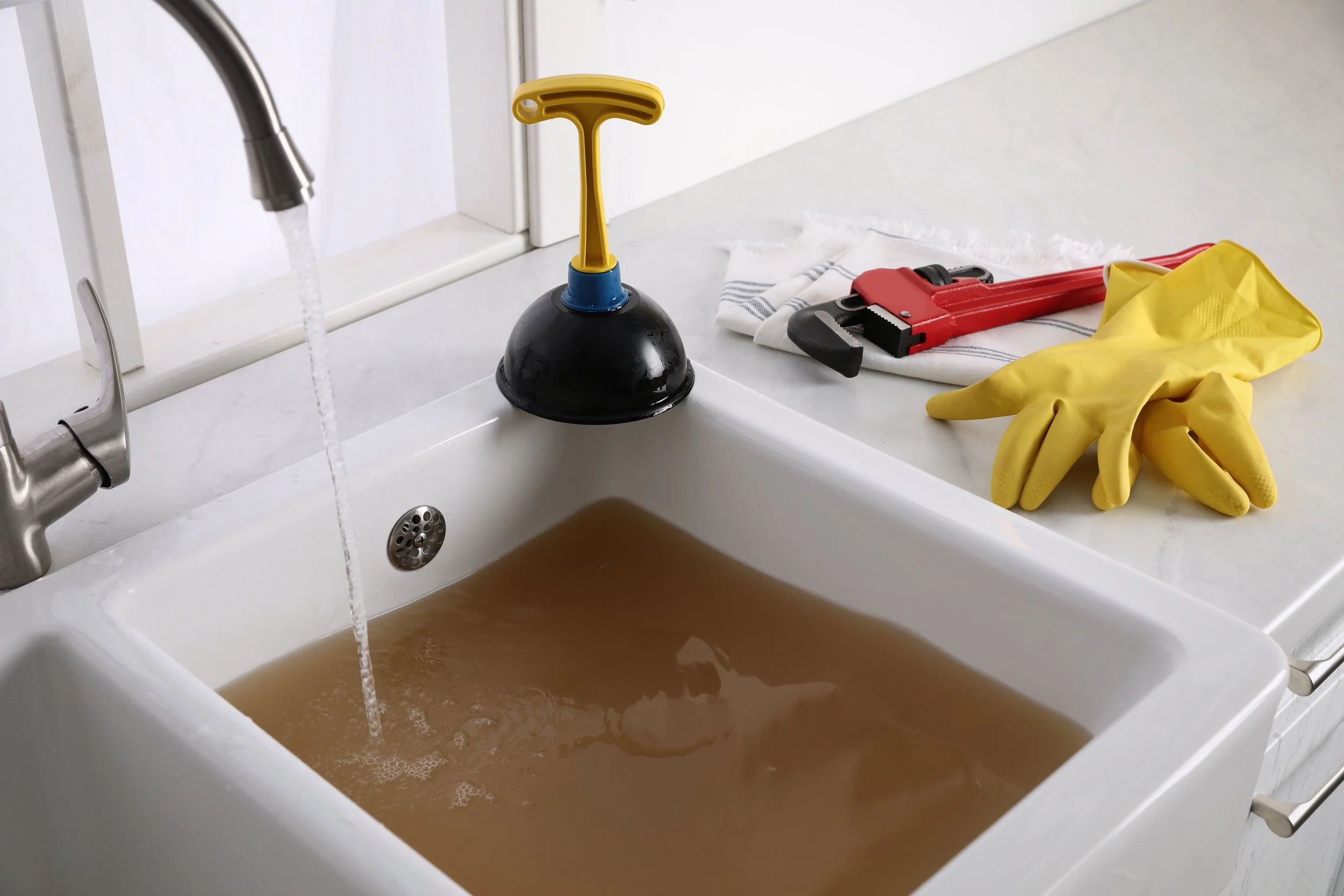 A kitchen sink filled with dirty water, with cleaning tools on the countertop including a plunger, rubber gloves, a wrench, a cloth, and a small black and red item.