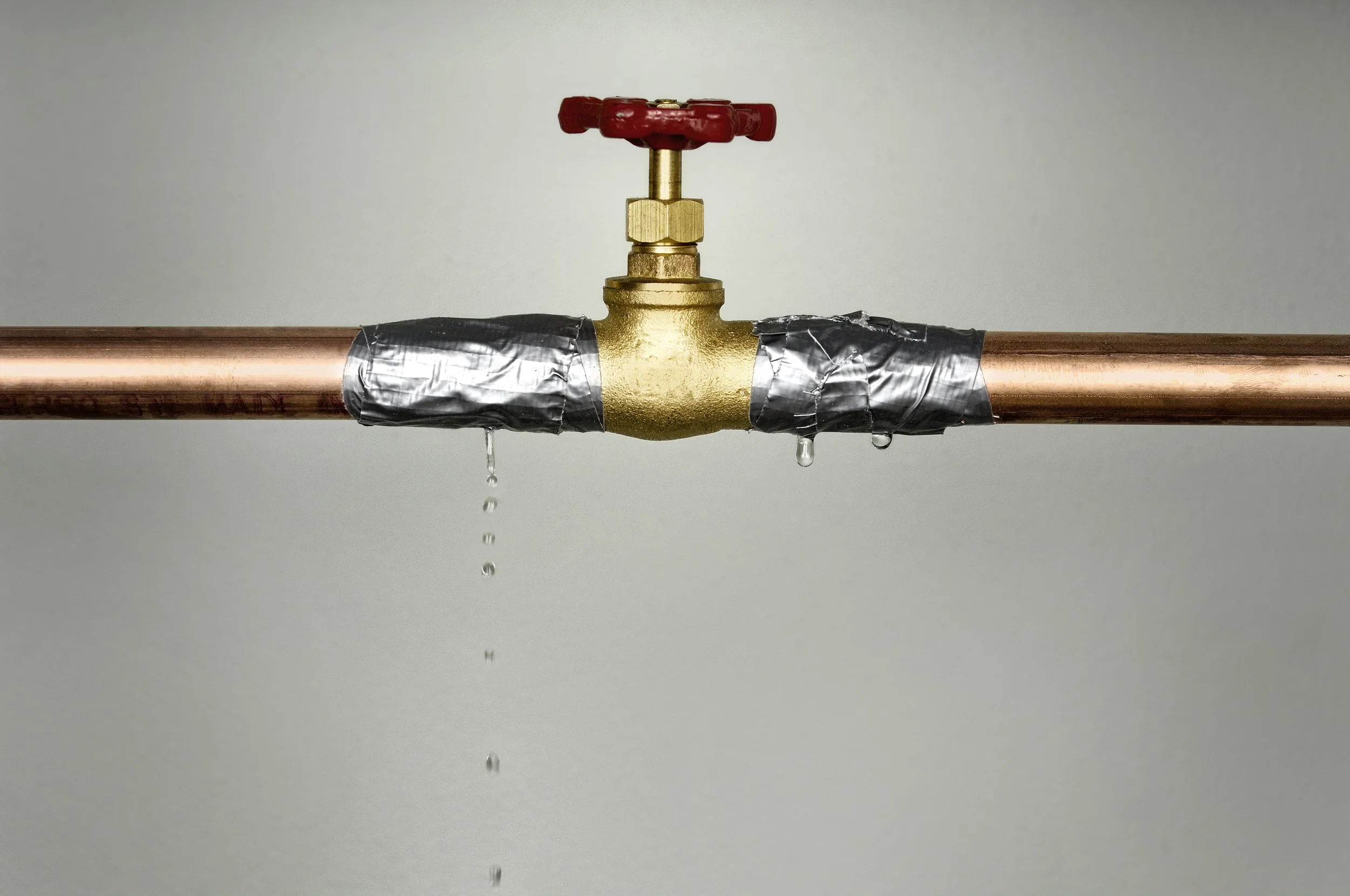 Water dripping from a black tape-sealed pipe connected to a gold-colored valve with a red handle.
