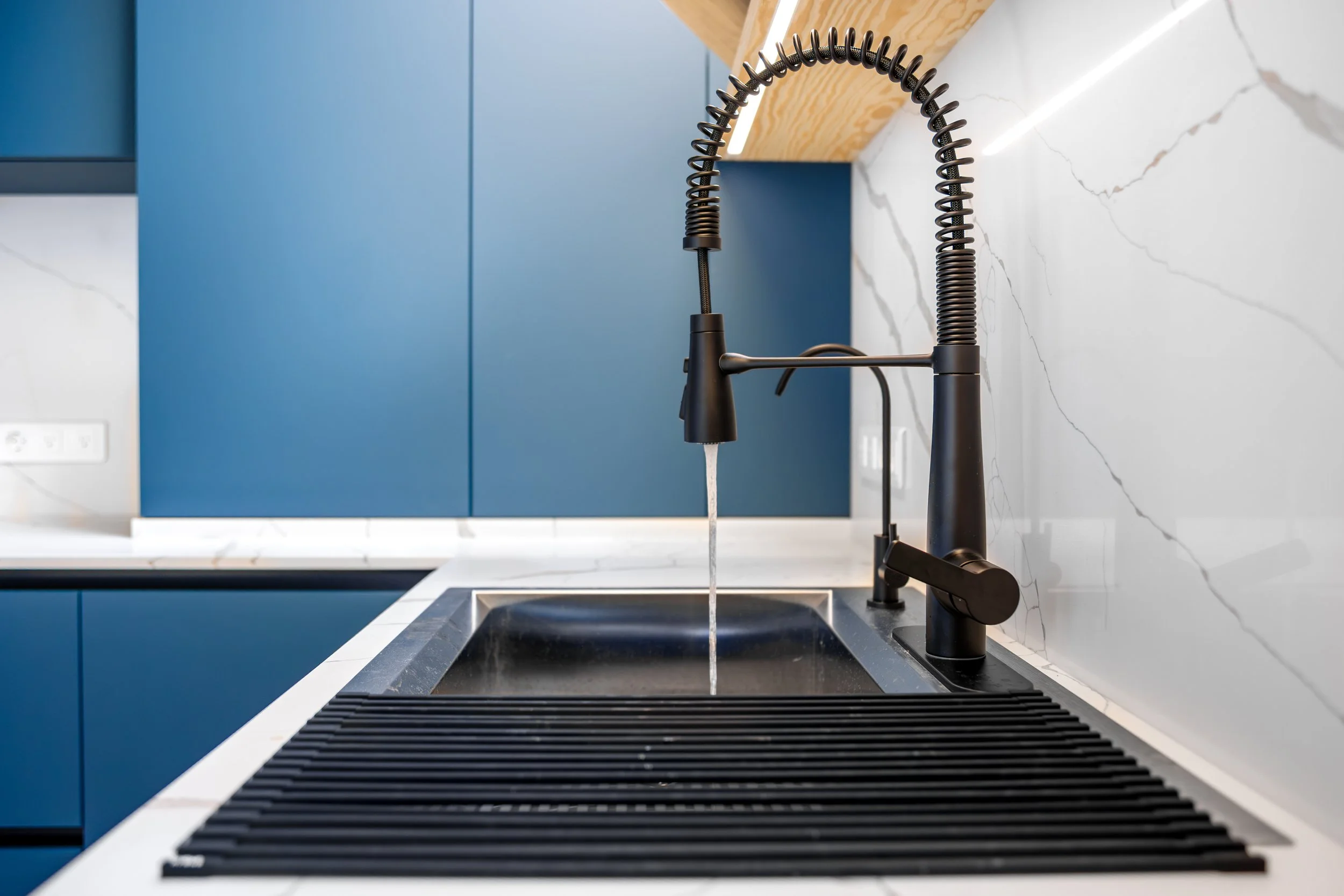 Modern kitchen sink with black faucet and black draining board, white marble backsplash, and blue cabinetry.