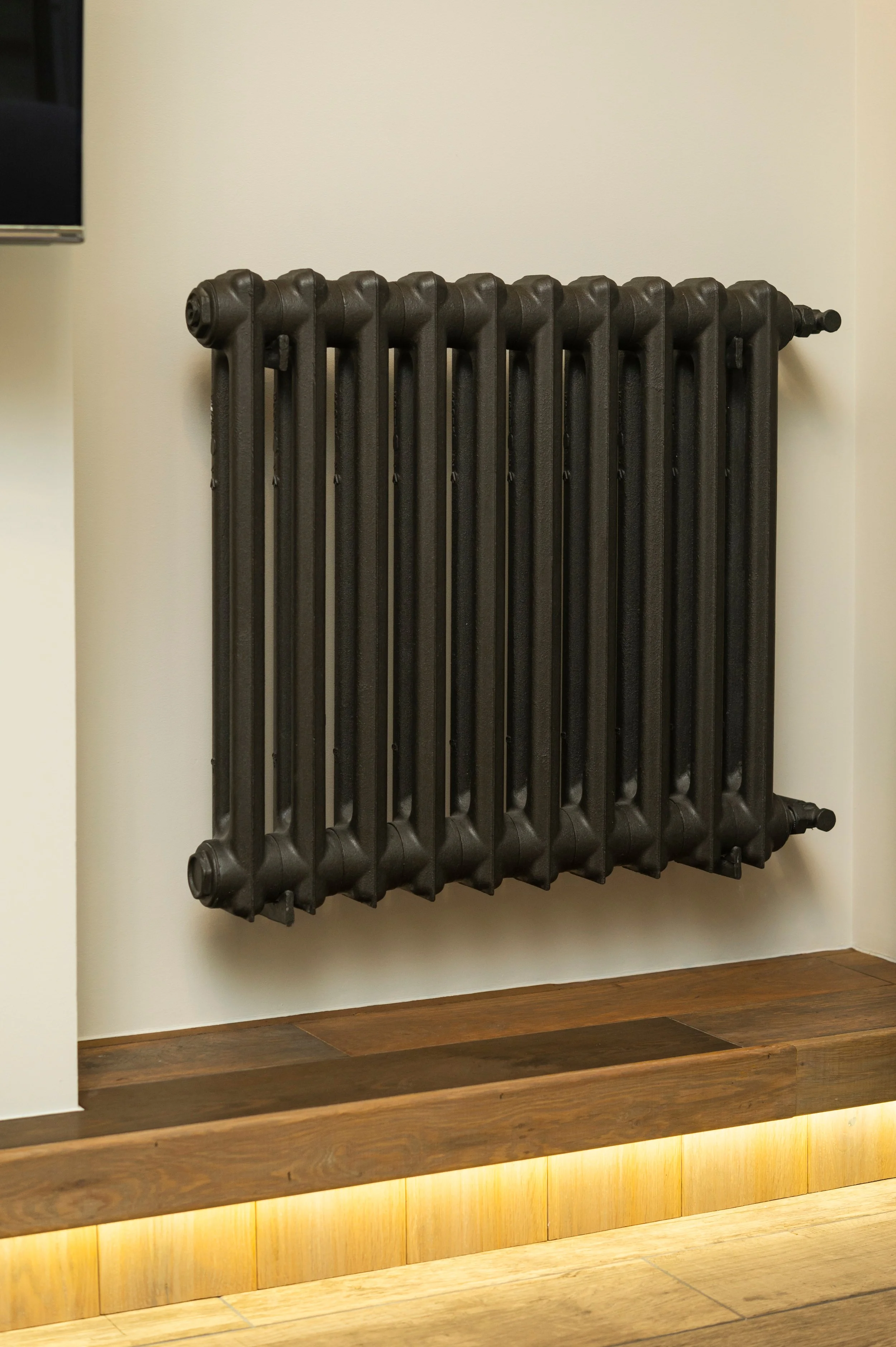Black cast iron radiator mounted on a beige wall with a wooden bench below and warm lighting along the bottom.