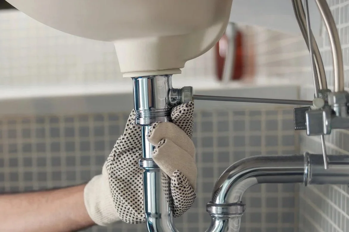 A person wearing a beige work glove is tightening a plumbing connector under a sink.